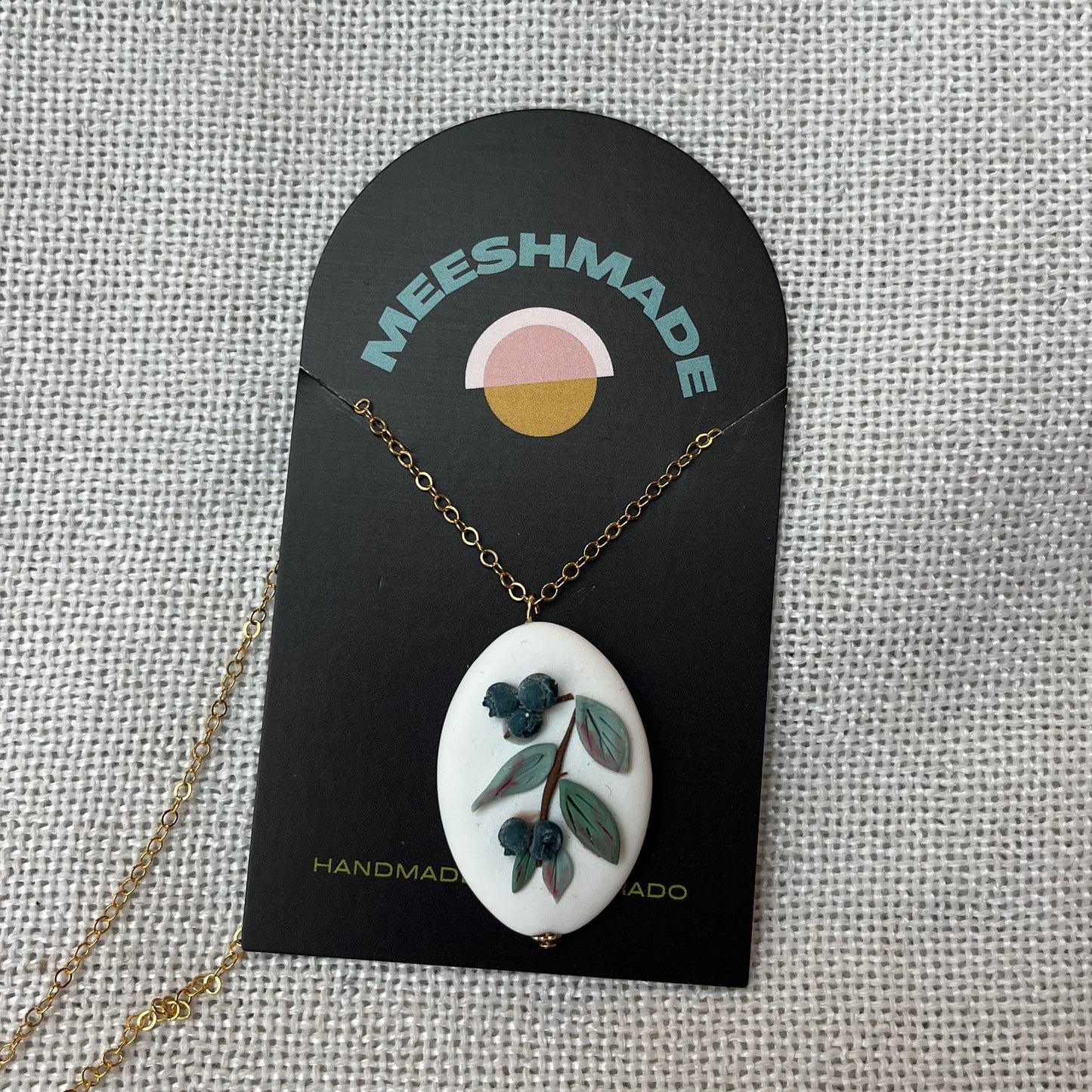 polymer clay necklace with blueberries design on white arch clay backdrop with gold chain, secured on branded jewelry card and resting on fabric background