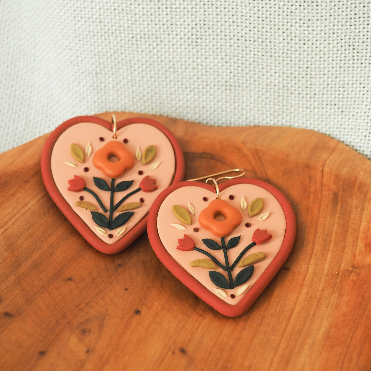 Folk style floral earrings in warm terra cotta tones on a soft heart-shaped background with gold filled fish hook hardware, set on a wooden backdrop