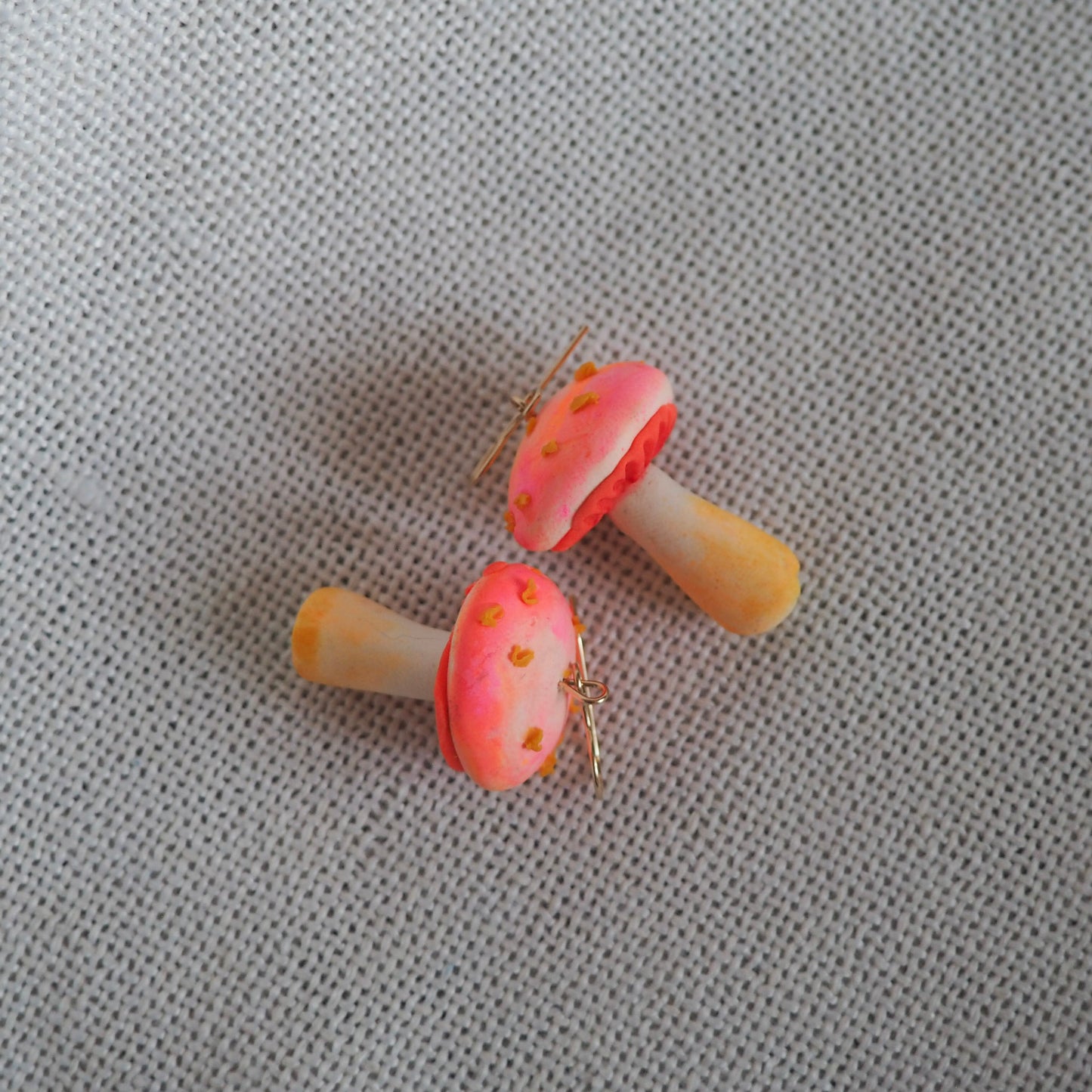 Pair of polymer clay earrings resembling small mushrooms with off white and yellow stems, electric coral/pink caps with yellow scales and gold hooks set on white textured background.