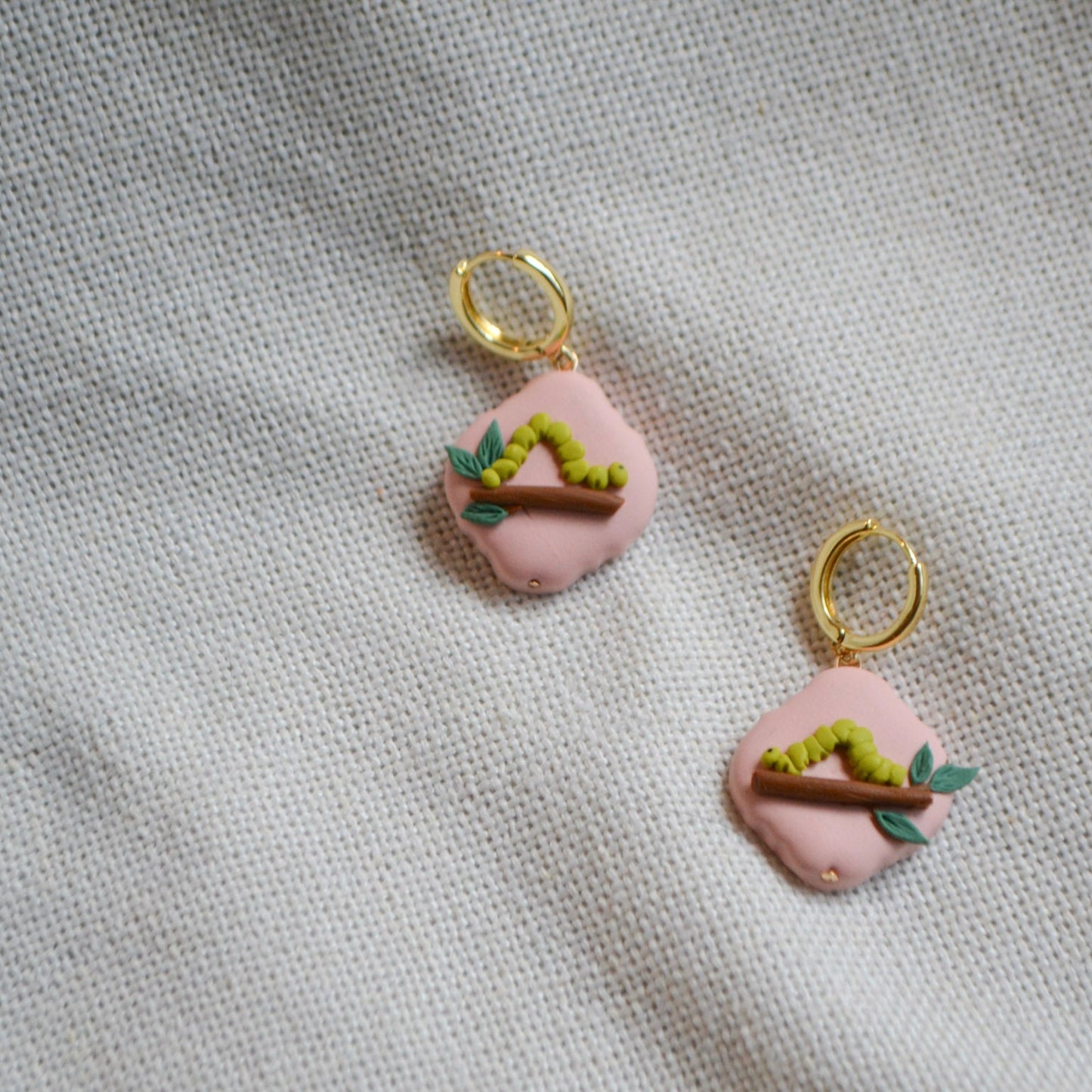 a pair of earrings with an inchworm on a leafy branch on a peach background with gold hoops
