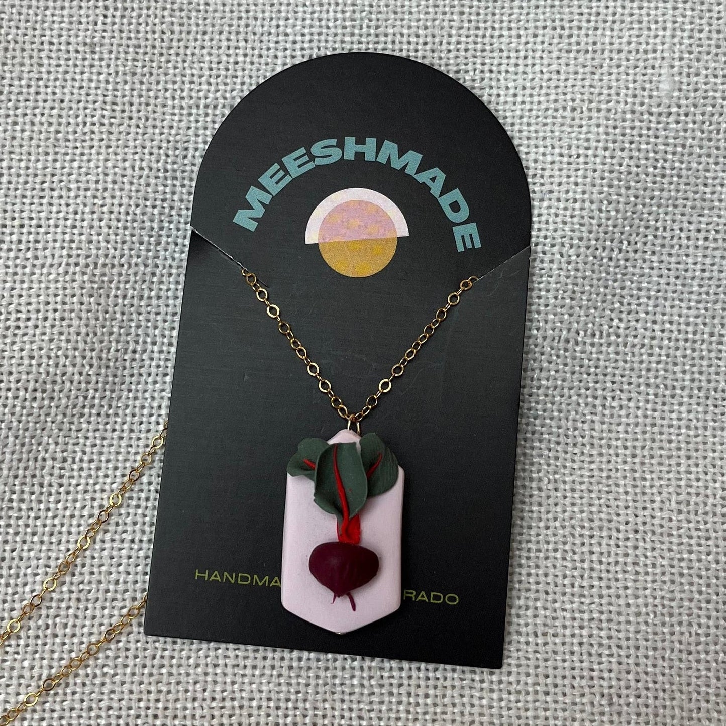 polymer clay necklace with beet root design on light pink geometric clay backdrop with gold chain, secured on branded jewelry card and resting on fabric background