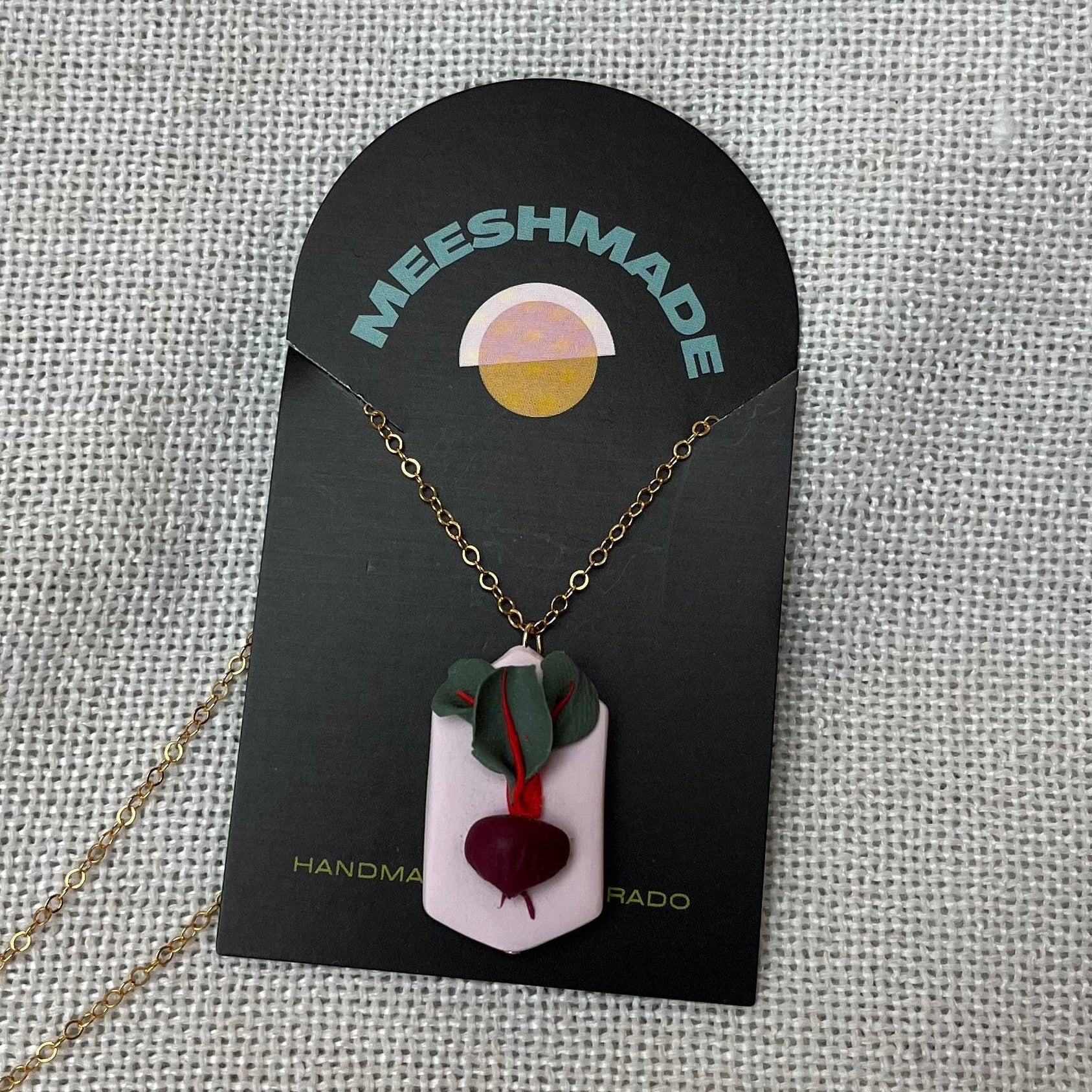 polymer clay necklace with beet root design on light pink geometric clay backdrop with gold chain, secured on branded jewelry card and resting on fabric background