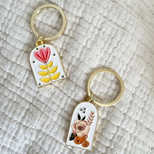 two white enamel keychains with floral design and gold ring on a cream fabric background. left side has pink and yellow flower, right lower keychain has cream and orange design
