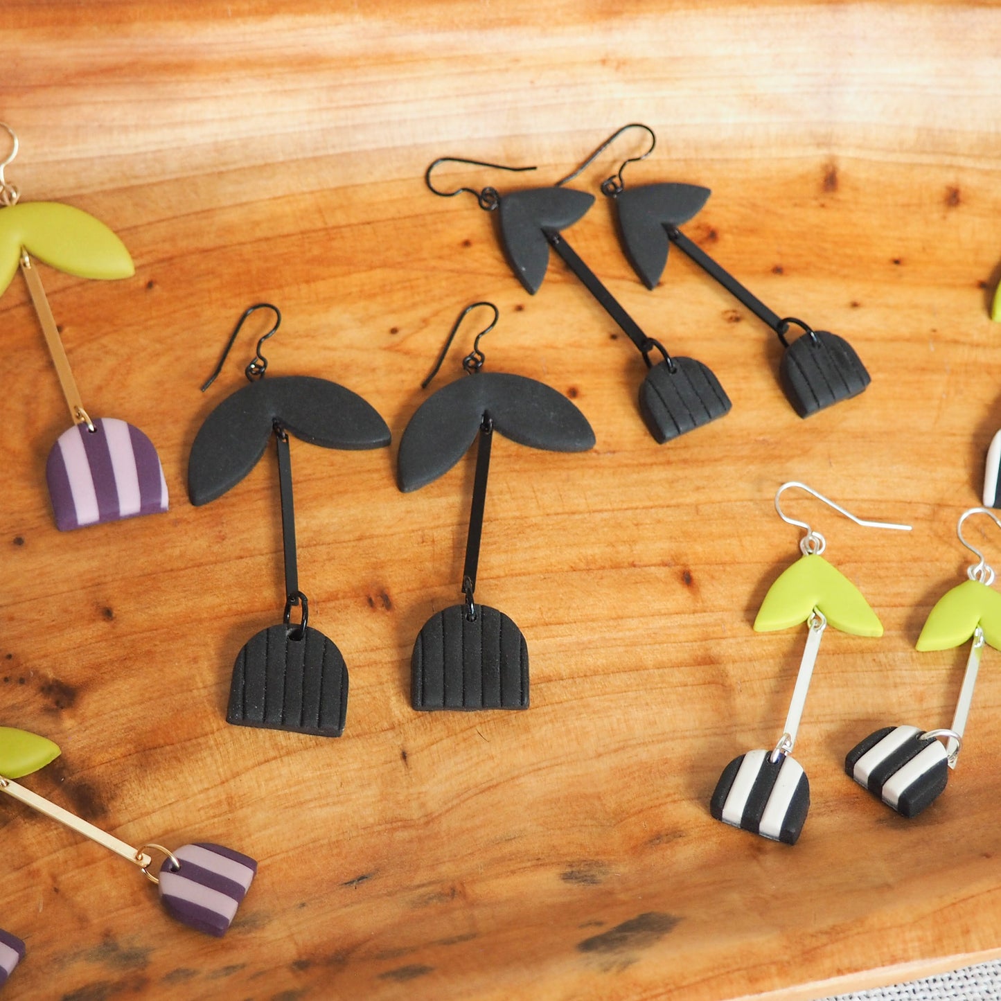 A selection of handmade polymer clay dangle earrings arranged on a wooden board. The earrings feature various designs including black palm tree silhouettes, black and white striped hearts, and lime green and purple striped hearts, some with black hooks and others with silver-tone hooks.