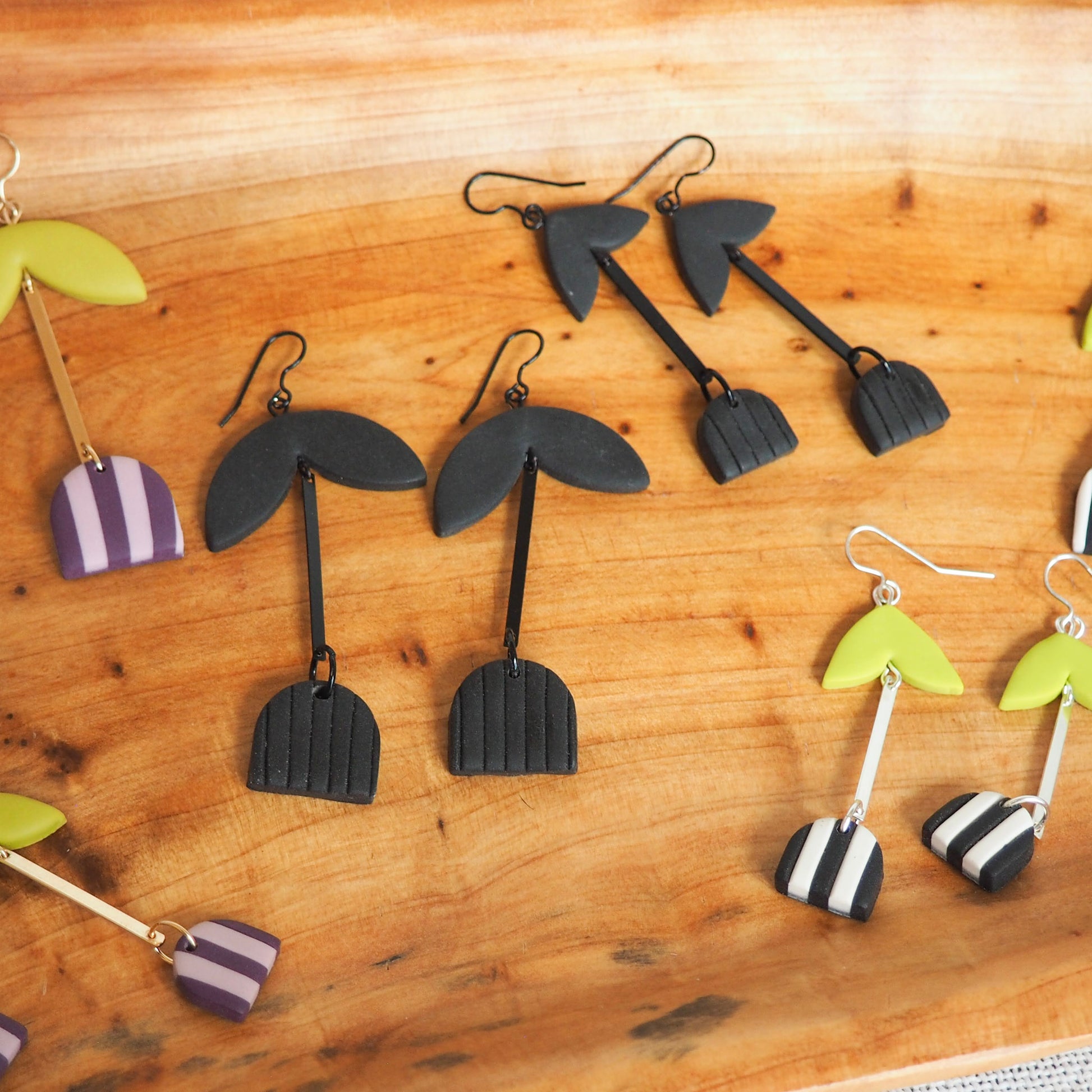 A selection of handmade polymer clay dangle earrings arranged on a wooden board. The earrings feature various designs including black palm tree silhouettes, black and white striped hearts, and lime green and purple striped hearts, some with black hooks and others with silver-tone hooks.