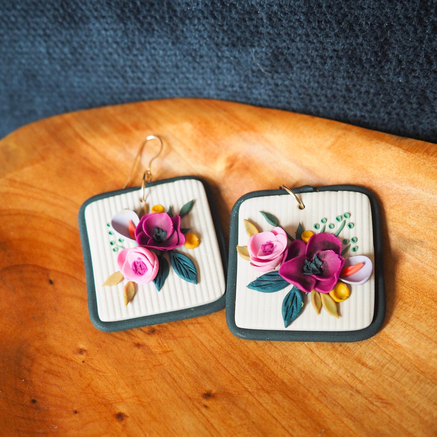 Polymer clay earrings with magenta, light pink, lavender florals on an off white background and a blue/gray border with gold hooks. Set on a wood and blue textured background. 