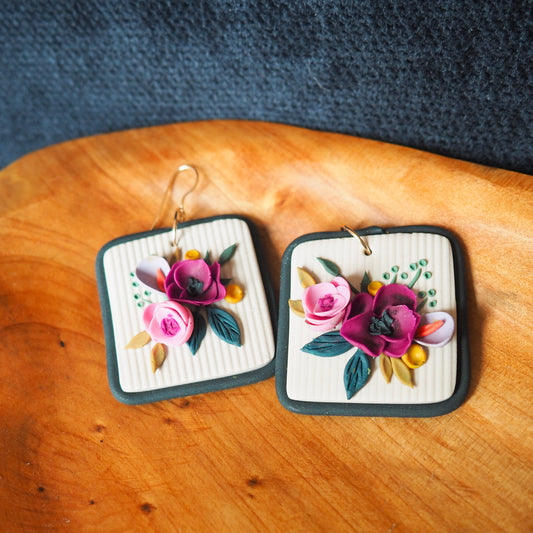Polymer clay earrings with magenta, light pink, lavender florals on an off white background and a blue/gray border with gold hooks. Set on a wood and blue textured background. 
