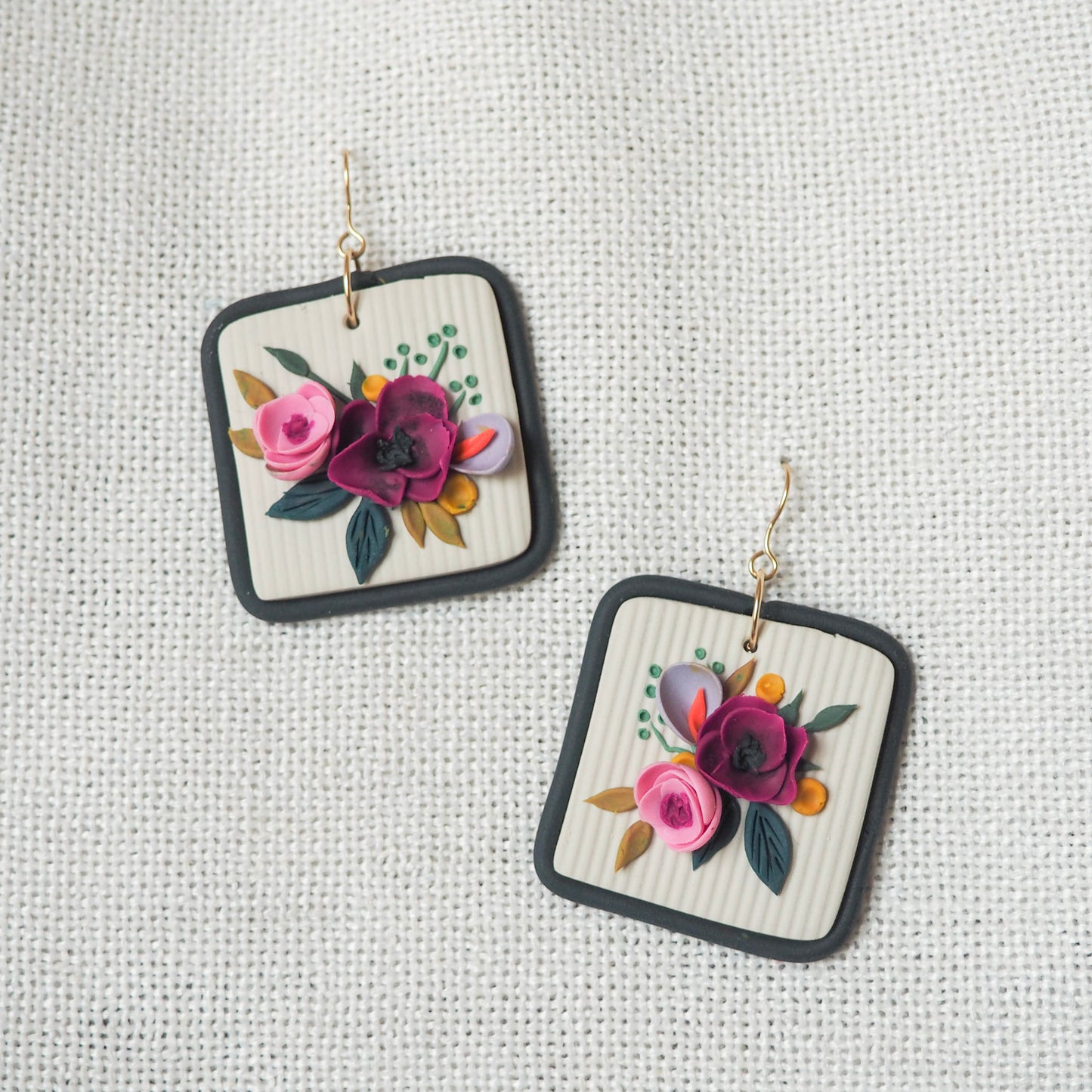 Polymer clay earrings with magenta, light pink, lavender florals on an off white background and a blue/gray border with gold hooks. Set on a white textured background. 