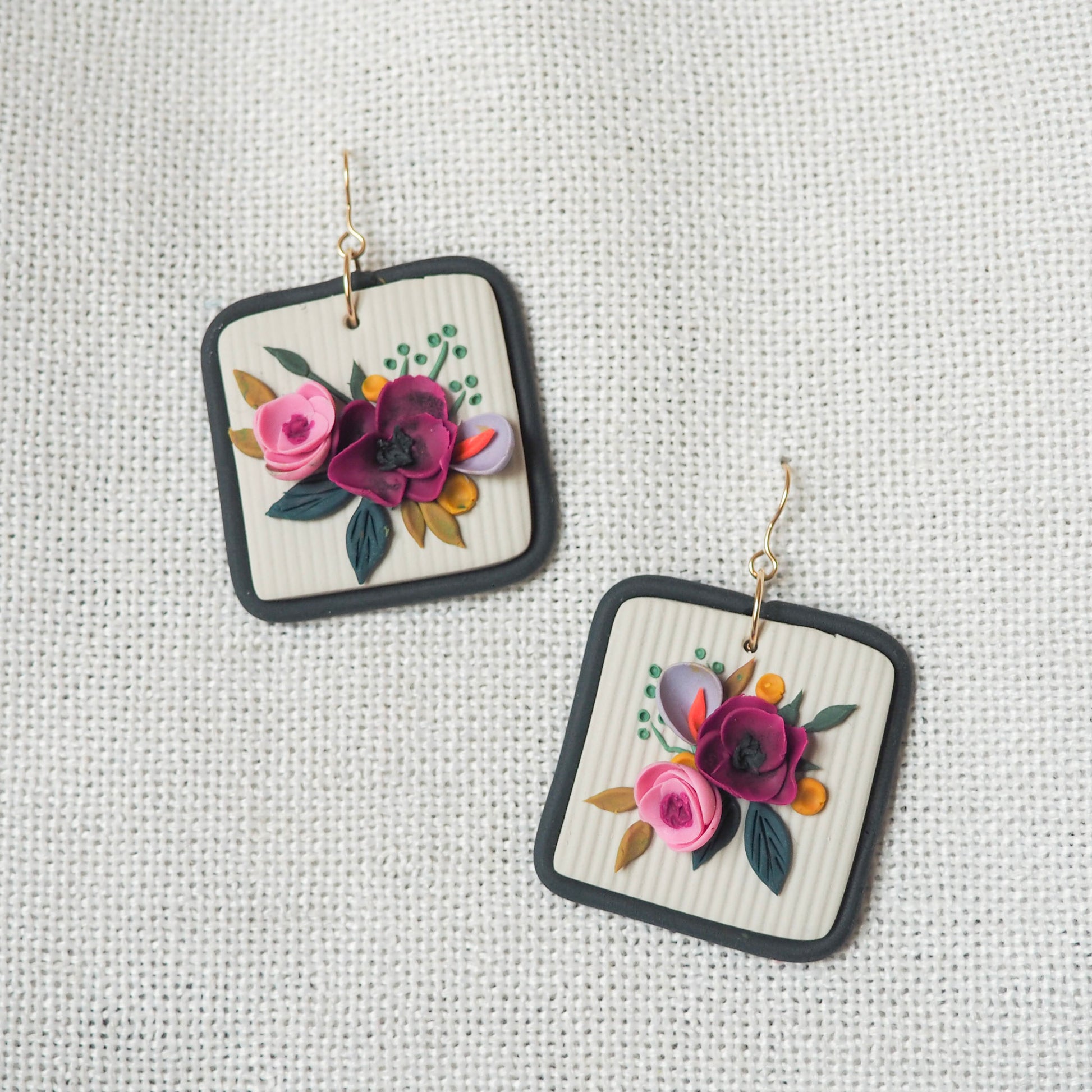 Polymer clay earrings with magenta, light pink, lavender florals on an off white background and a blue/gray border with gold hooks. Set on a white textured background. 