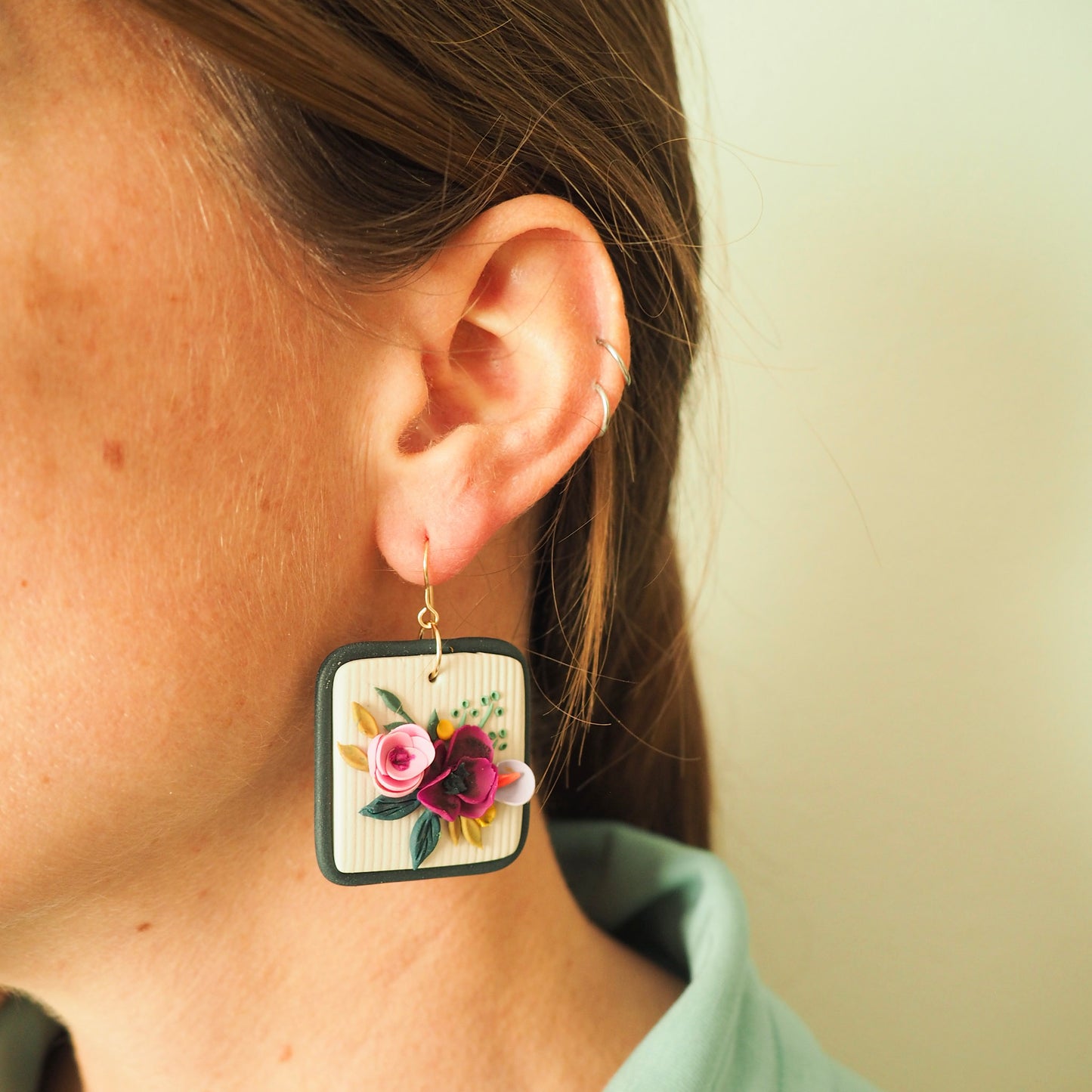 Model wearing polymer clay earrings with magenta, light pink, lavender florals on an off white background and a blue/gray border with gold hooks. 
