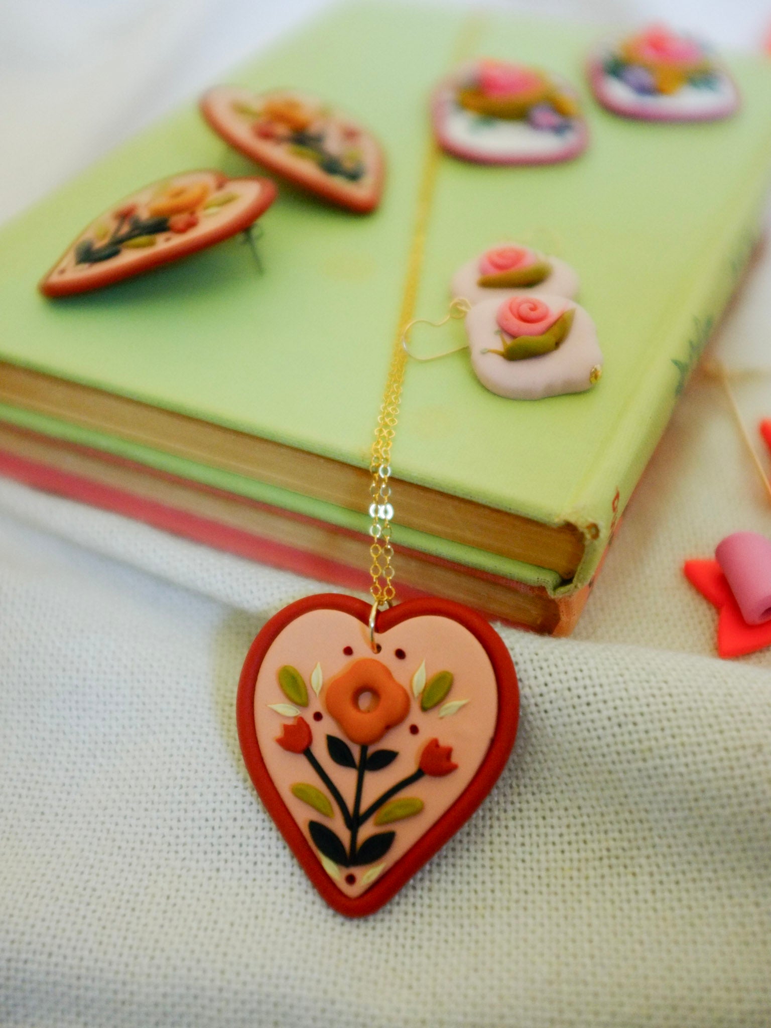 Folk style floral necklace with warm terra cotta colors on a soft heart-shaped background with gold filled chain, set on a light green book and fabric backdrop with other earrings in the background