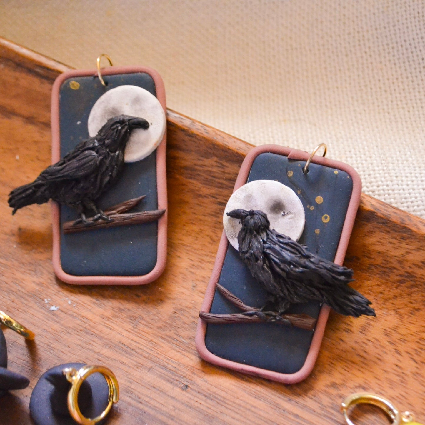 A pair of handmade gothic-style earrings featuring black crows perched in front of a white full moon on a dark slate-blue background, set in a dusty pink rectangular frame with gold hooks, displayed on a wooden surface.