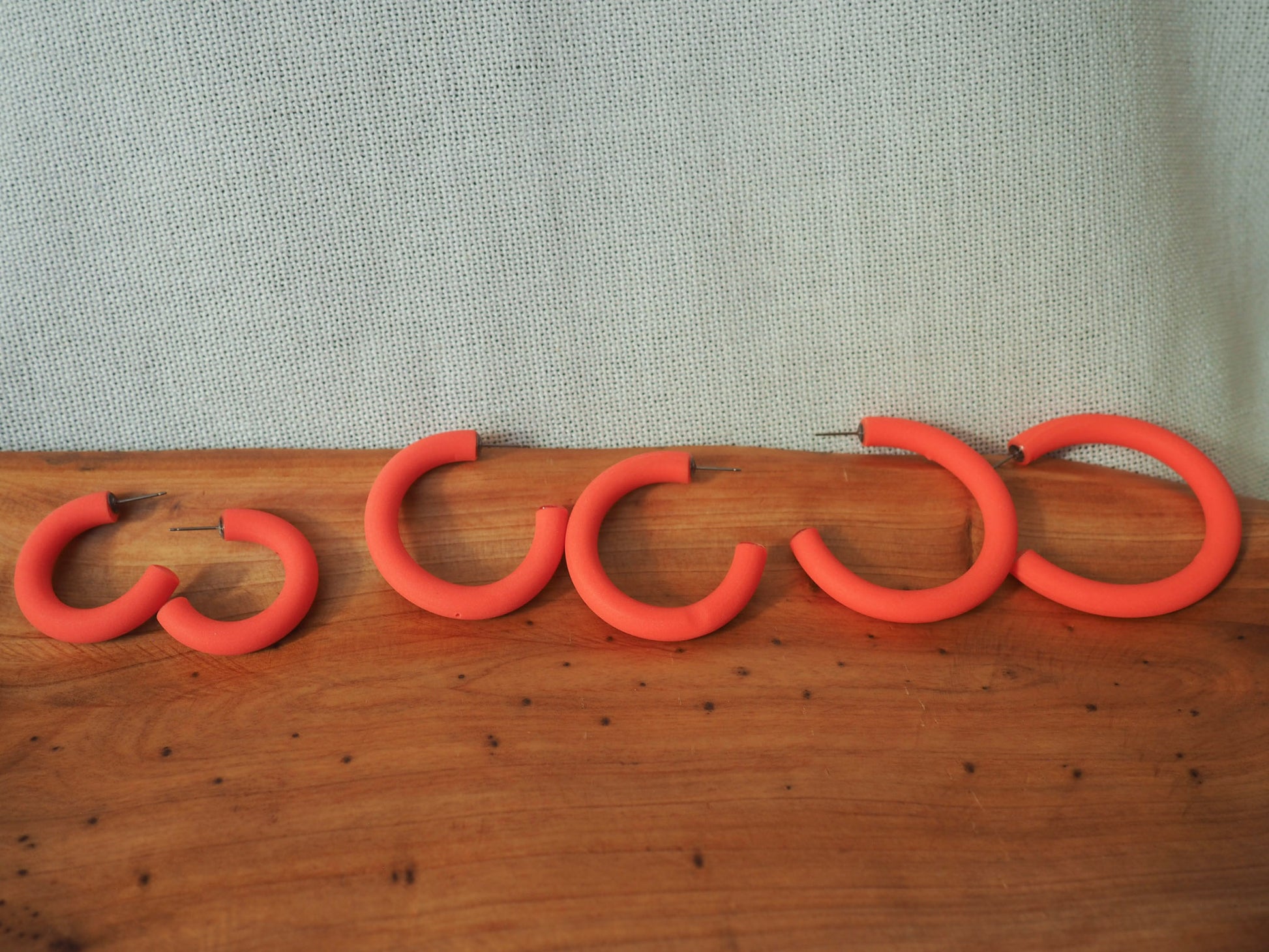 coral hoop earrings lined up from small to medium to large sizes on a wooden and textured fabric background
