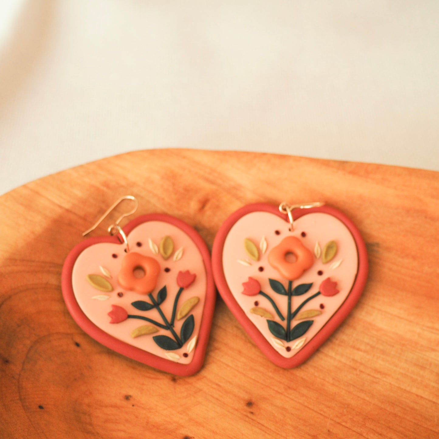 Folk style floral earrings in warm terra cotta tones on a soft heart-shaped background with gold filled fish hook hardware, set on a wooden backdrop