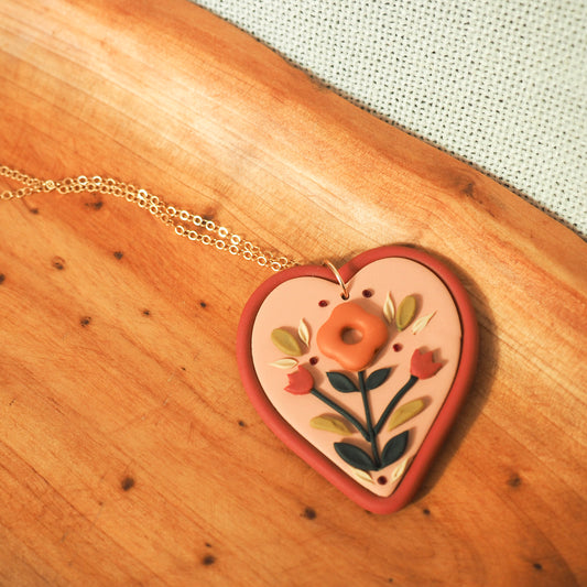 Folk style floral necklace with warm terra cotta colors on a soft heart-shaped background with gold filled chain, set on a wooden backdrop