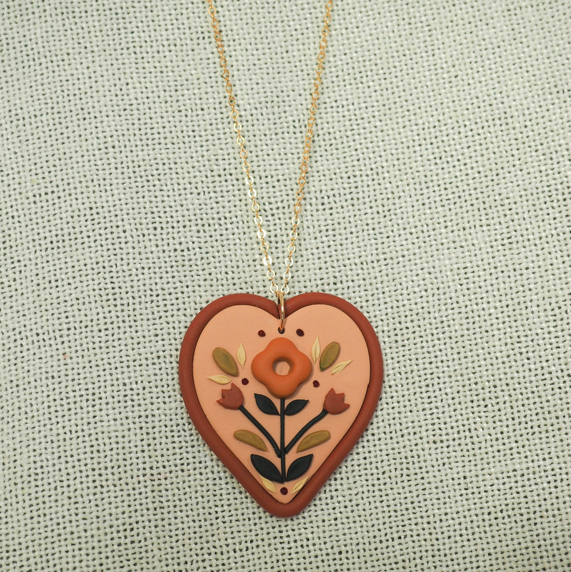 Folk style floral necklace with warm terra cotta colors on a soft heart-shaped background with gold filled chain, set on a textured cream fabric backdrop