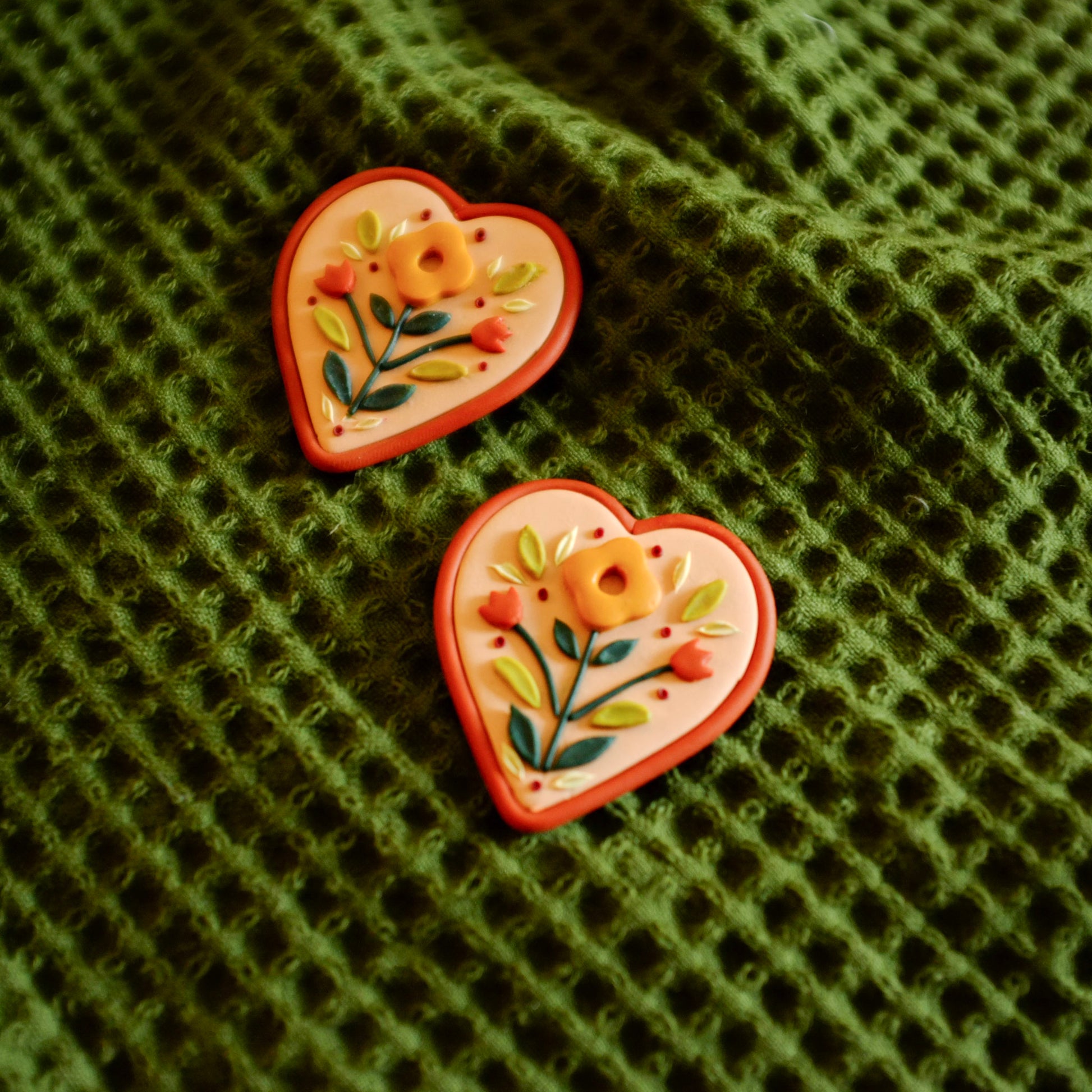 Folk style and heart shaped clay floral earrings with warm terra cotta colors, set on a dark green backdrop