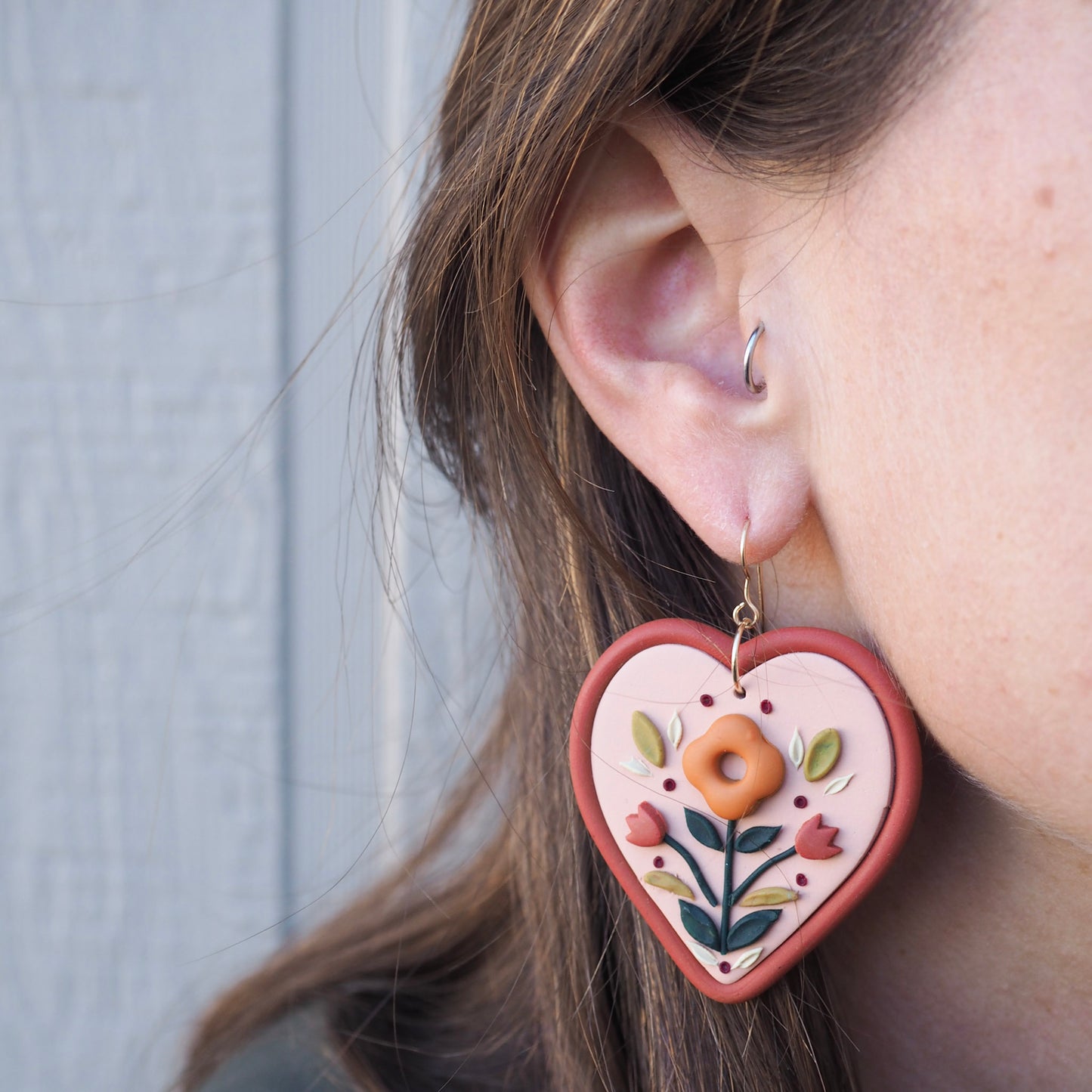 model wearing Folk style floral earrings in warm terra cotta tones on a soft heart-shaped background with gold filled fish hook hardware