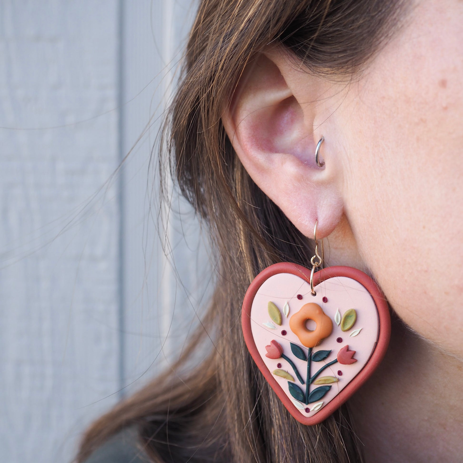model wearing Folk style floral earrings in warm terra cotta tones on a soft heart-shaped background with gold filled fish hook hardware