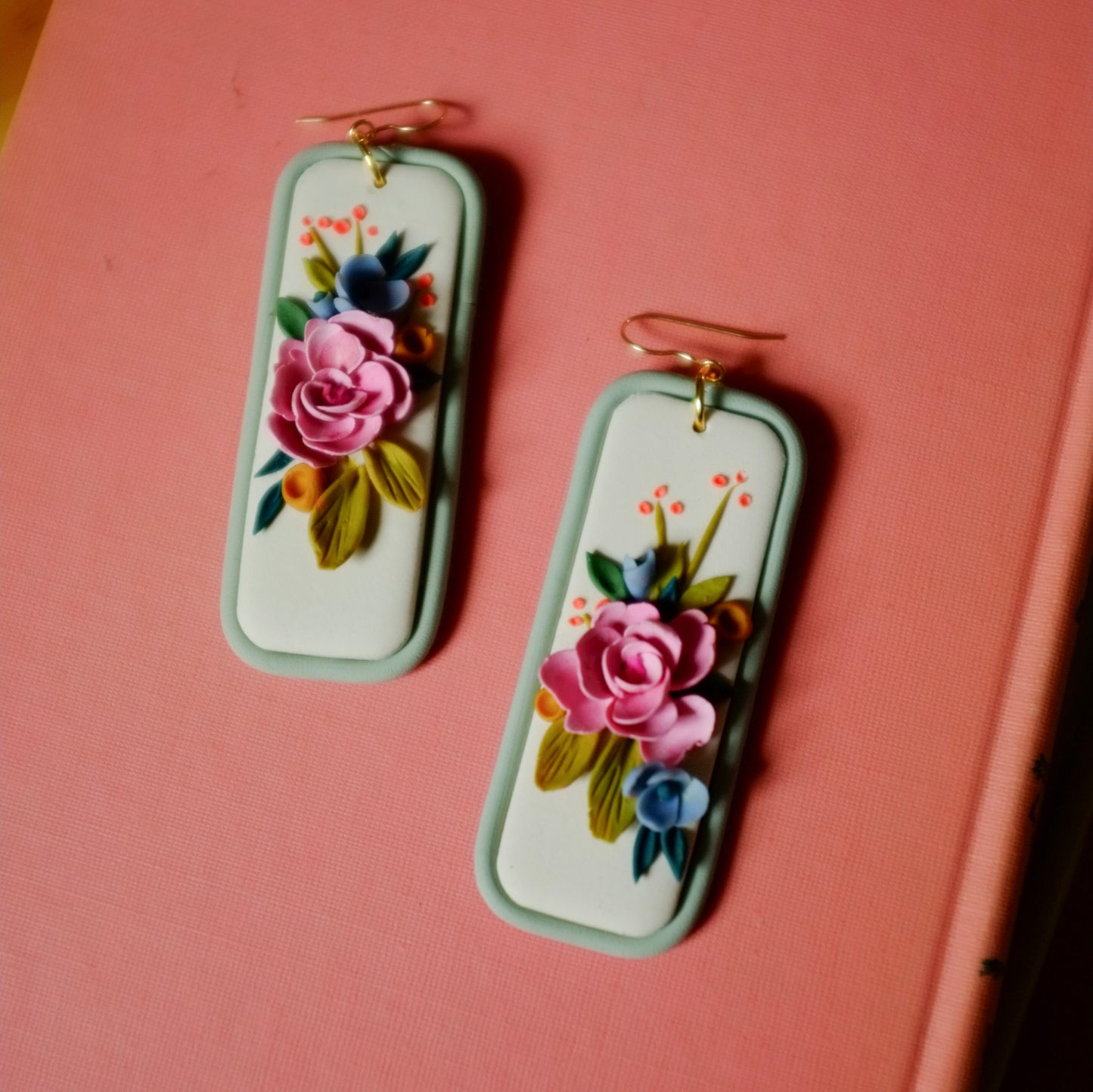 pink and blue florals earrings with orange and green accents on a light blue rectangle background with gold fish hooks, set on coral backdrop