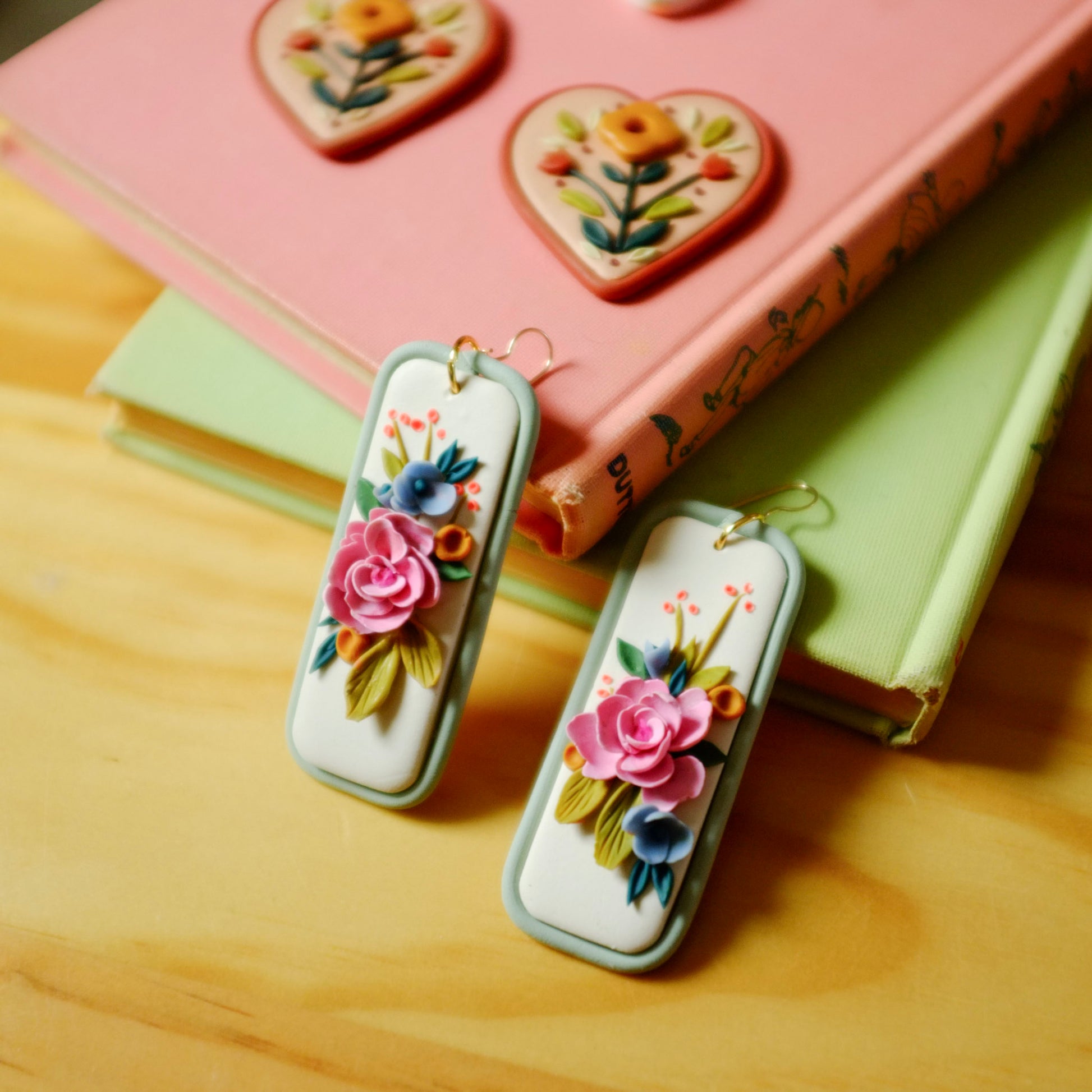 pink and blue florals earrings with orange and green accents on a light blue rectangle background with gold fish hooks, leaning against coral and light green books with wooden surface and other earrings in the background