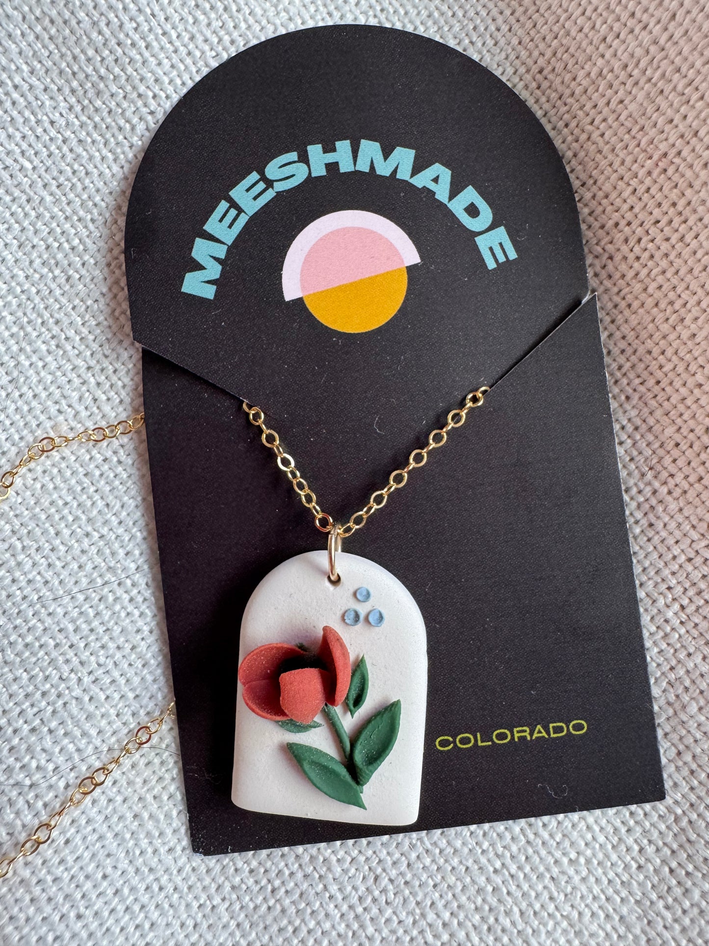 polymer clay necklace with red poppy flower design on white arch clay backdrop with gold chain, secured on branded jewelry card and resting on fabric background