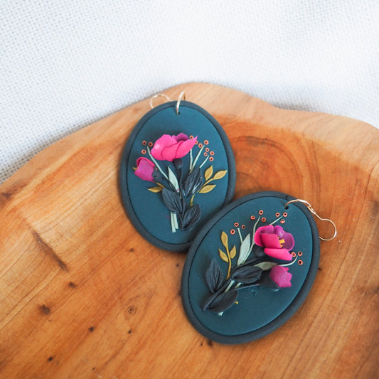 Polymer clay earrings with fuchsia floral on a dark blue background and green and orange accents with gold hooks. Set on a wood and white textured background. 