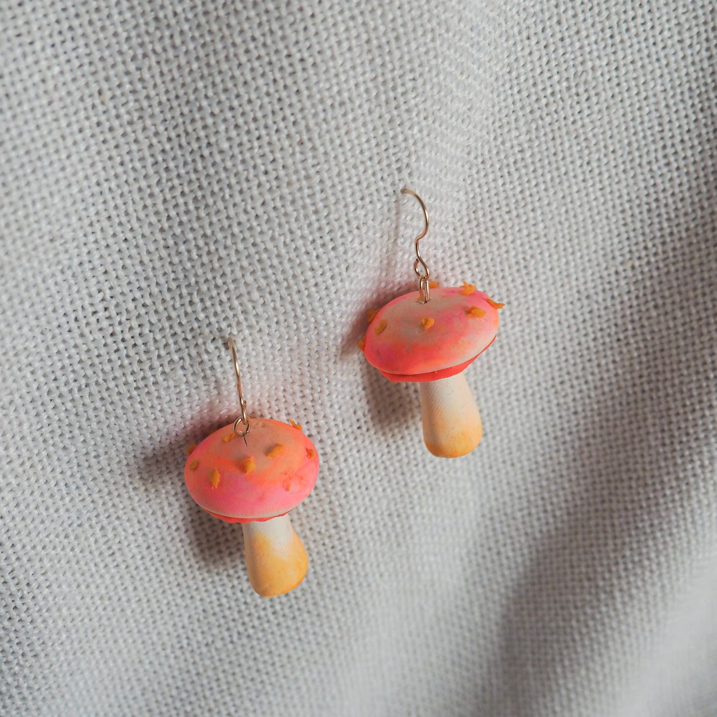 Pair of polymer clay earrings resembling small mushrooms with off white and yellow stems, electric coral/pink caps with yellow scales and gold hooks set on white textured background. 