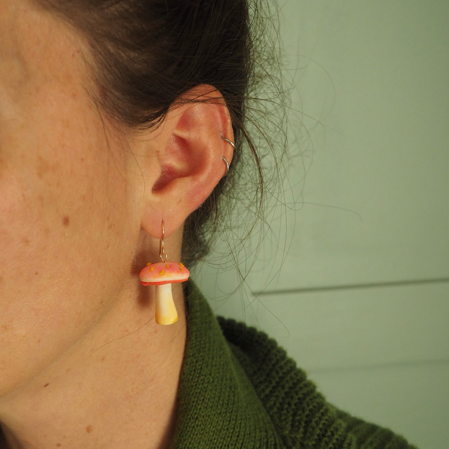 Model wearing a polymer clay earrings resembling a small mushroom with off white and yellow stem, electric coral/pink cap with yellow scales with a gold hook. 