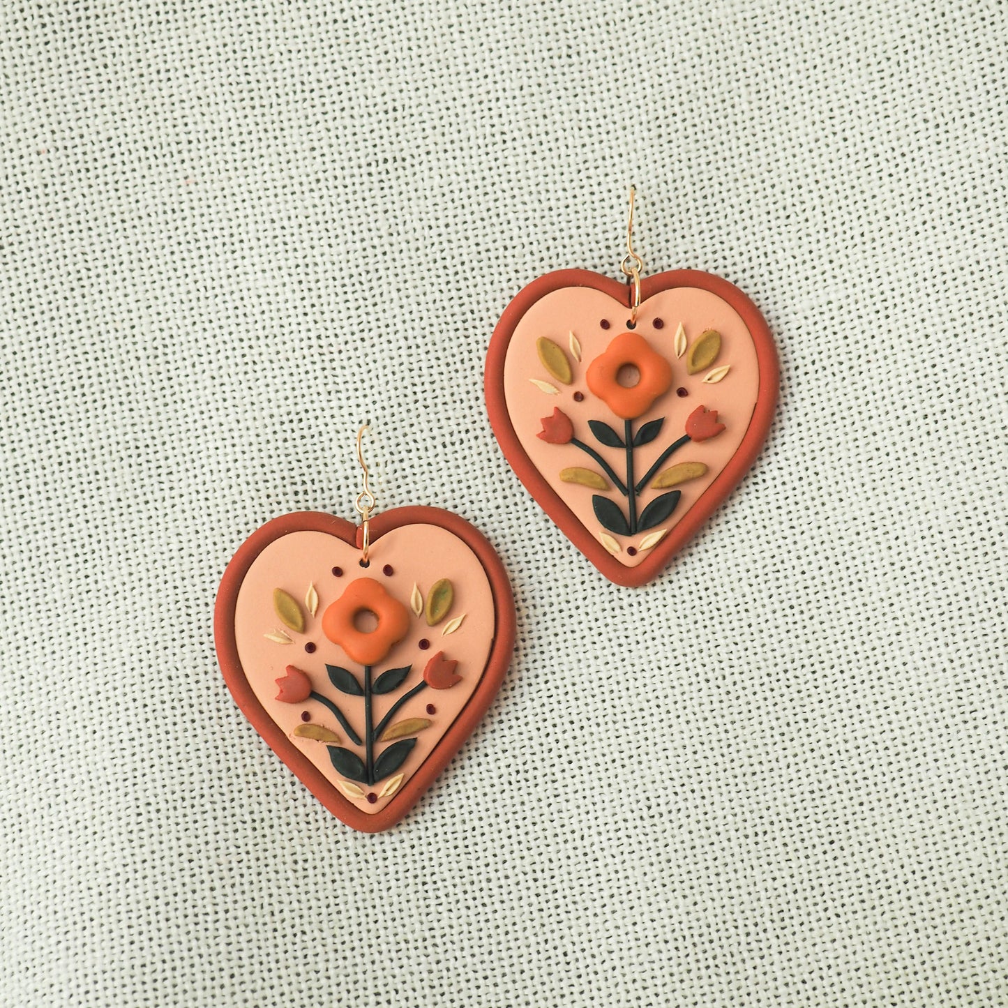 Folk style floral earrings in warm terra cotta tones on a soft heart-shaped background with gold filled fish hook hardware, set on a textured fabric backdrop