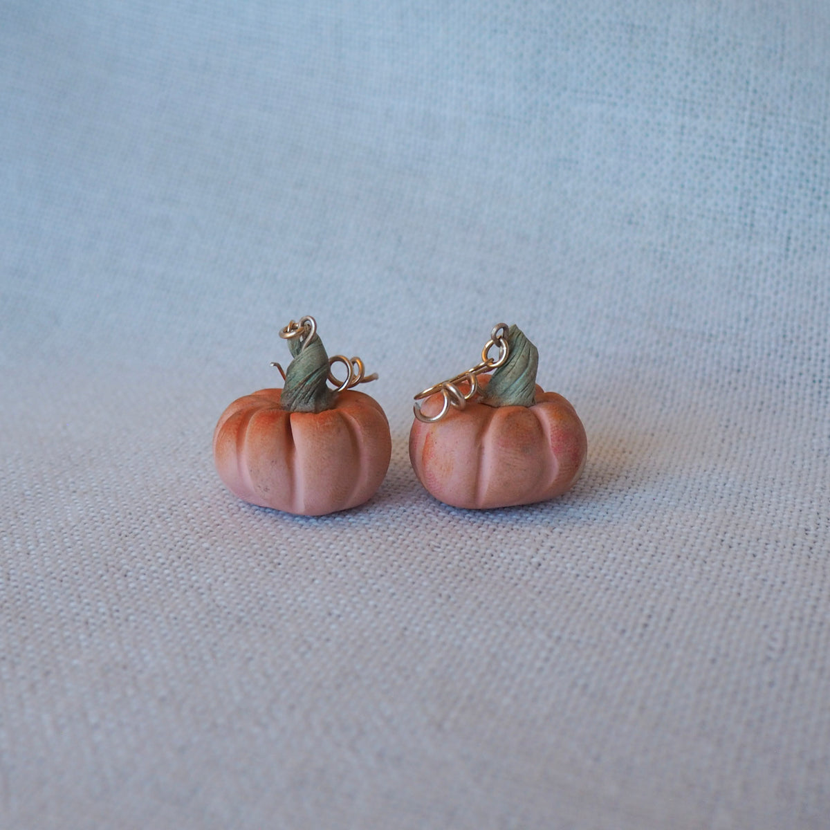 A pair of small pumpkin-shaped earrings with muted peach-orange bodies, green stems, and gold-tone hooks, displayed on a light gray textured fabric background.