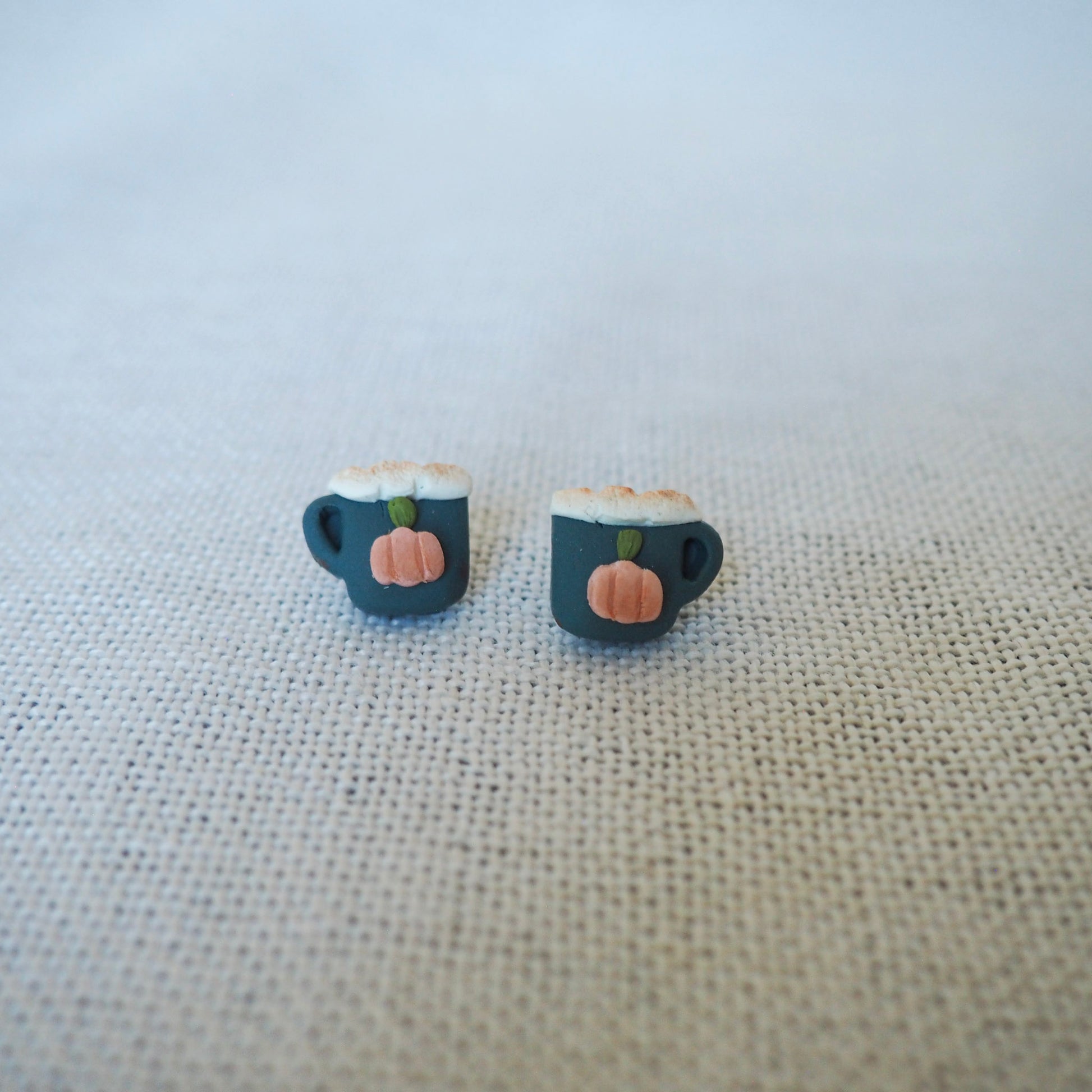 A pair of small stud earrings shaped like dark teal coffee mugs with white foam and a small orange pumpkin on the front, displayed on a textured beige fabric background.