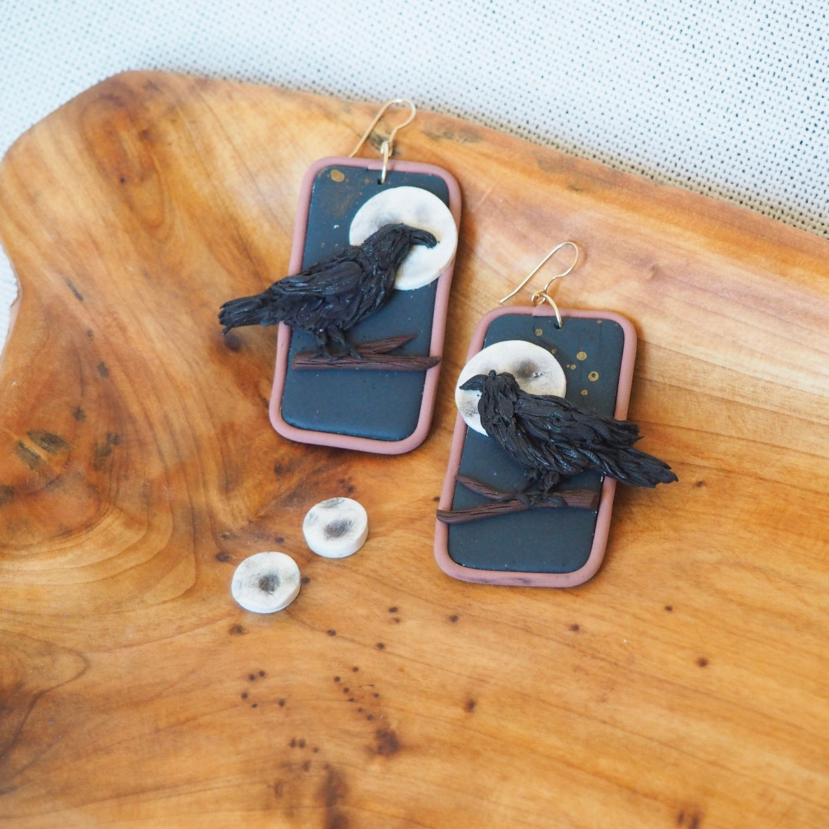 A pair of handmade rectangular earrings with black raven figurines perched on branches against a dark blue-gray background, featuring a pale cream moon and gold-toned hooks, displayed on a wooden board.

A pair of moon stud earrings sits below the raven earrings.