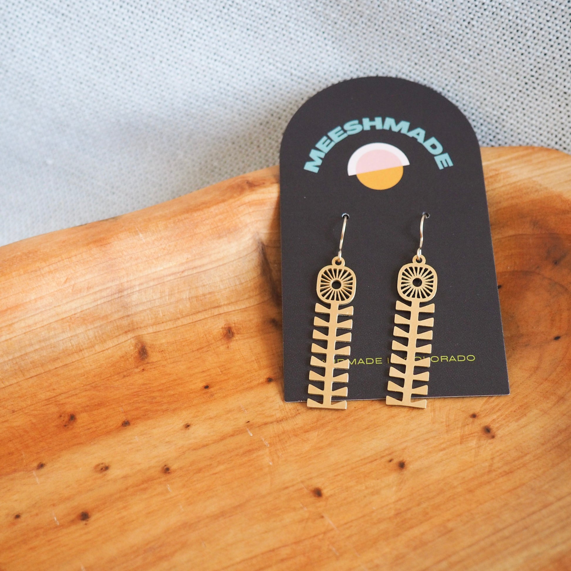folk style flower earrings made from bass with fish hooks on a black jewelry card set in a wooden tray on a white textured background 