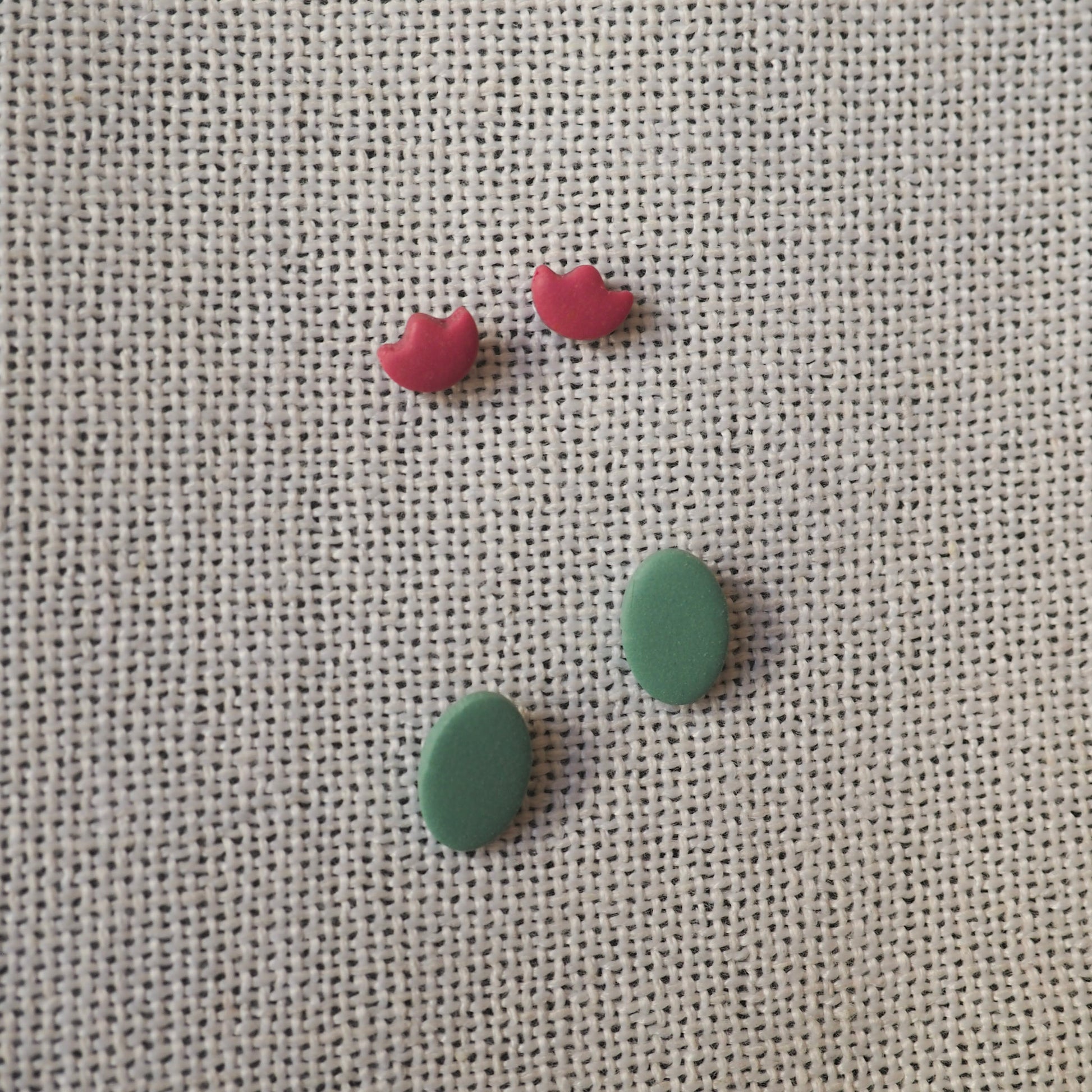 Two sets of stud polymer clay earrings, with light cranberry tulip earrings and blue/green oval earrings set on white textured backdrop 