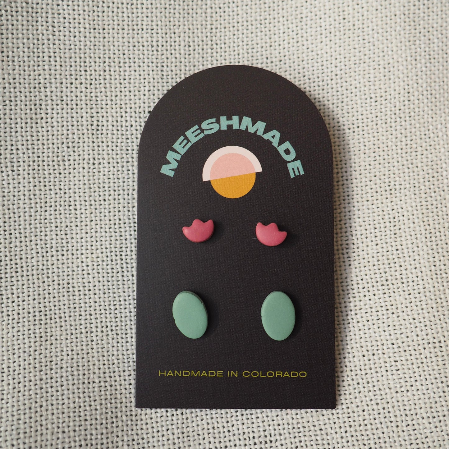 Two sets of stud polymer clay earrings, with light cranberry tulip earrings and blue/green ovals earrings on black card set on white textured backdrop 