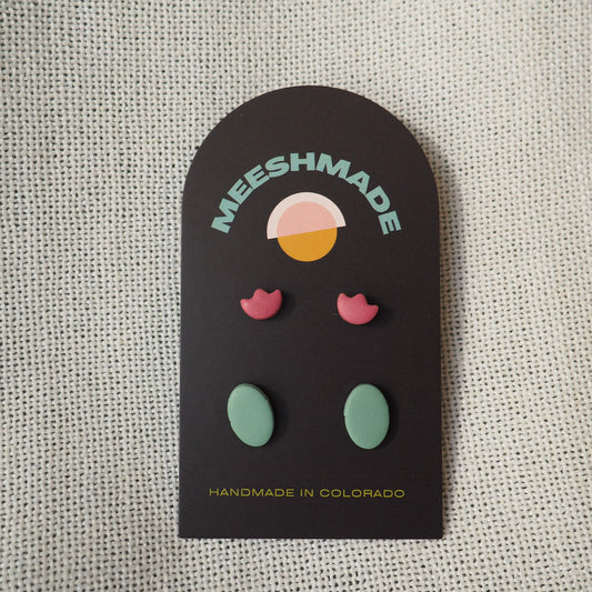 Two sets of stud polymer clay earrings, with light cranberry tulip earrings and blue/green ovals earrings on black card set on white textured backdrop 