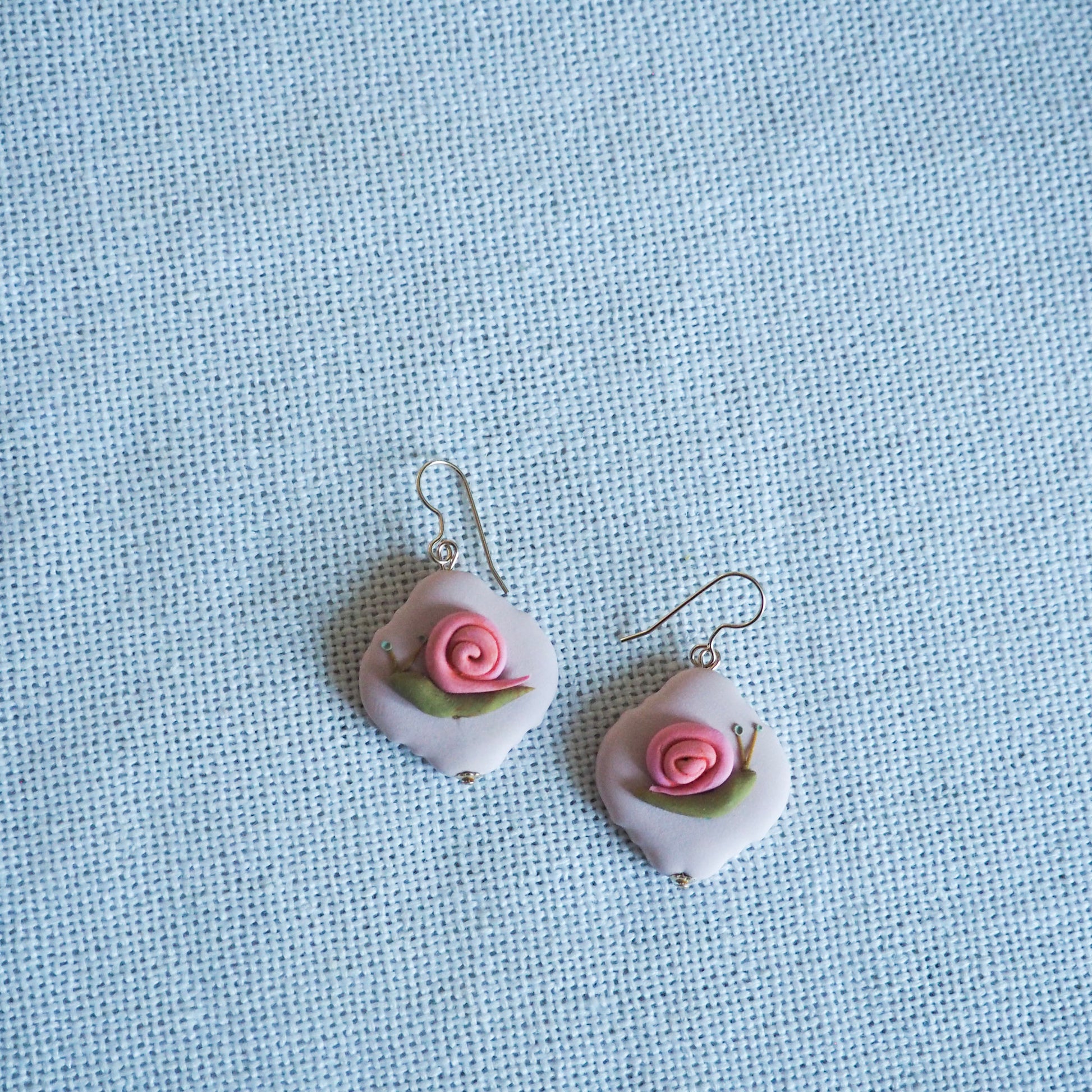 polymer clay earrings with gold hooks with green and pink snail on pink translucent background, set on a white fabric background