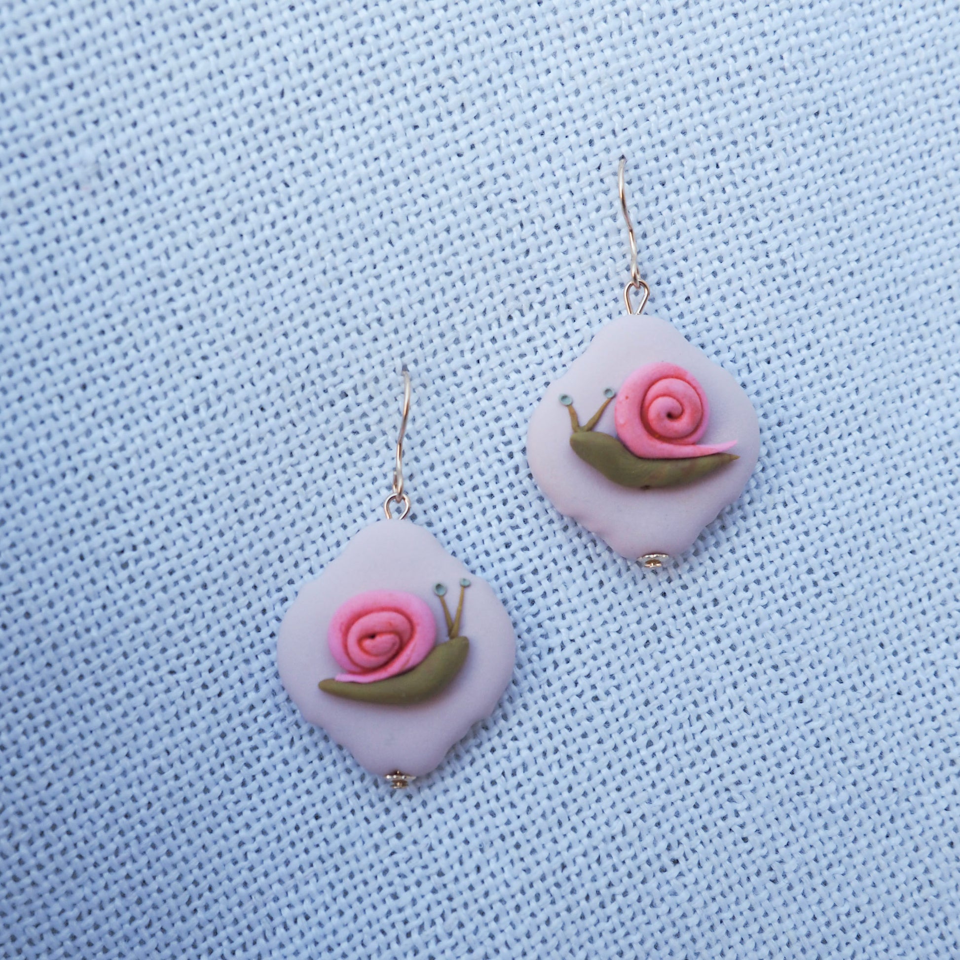 polymer clay earrings with gold hooks with green and pink snail on pink translucent background, set on a white fabric background