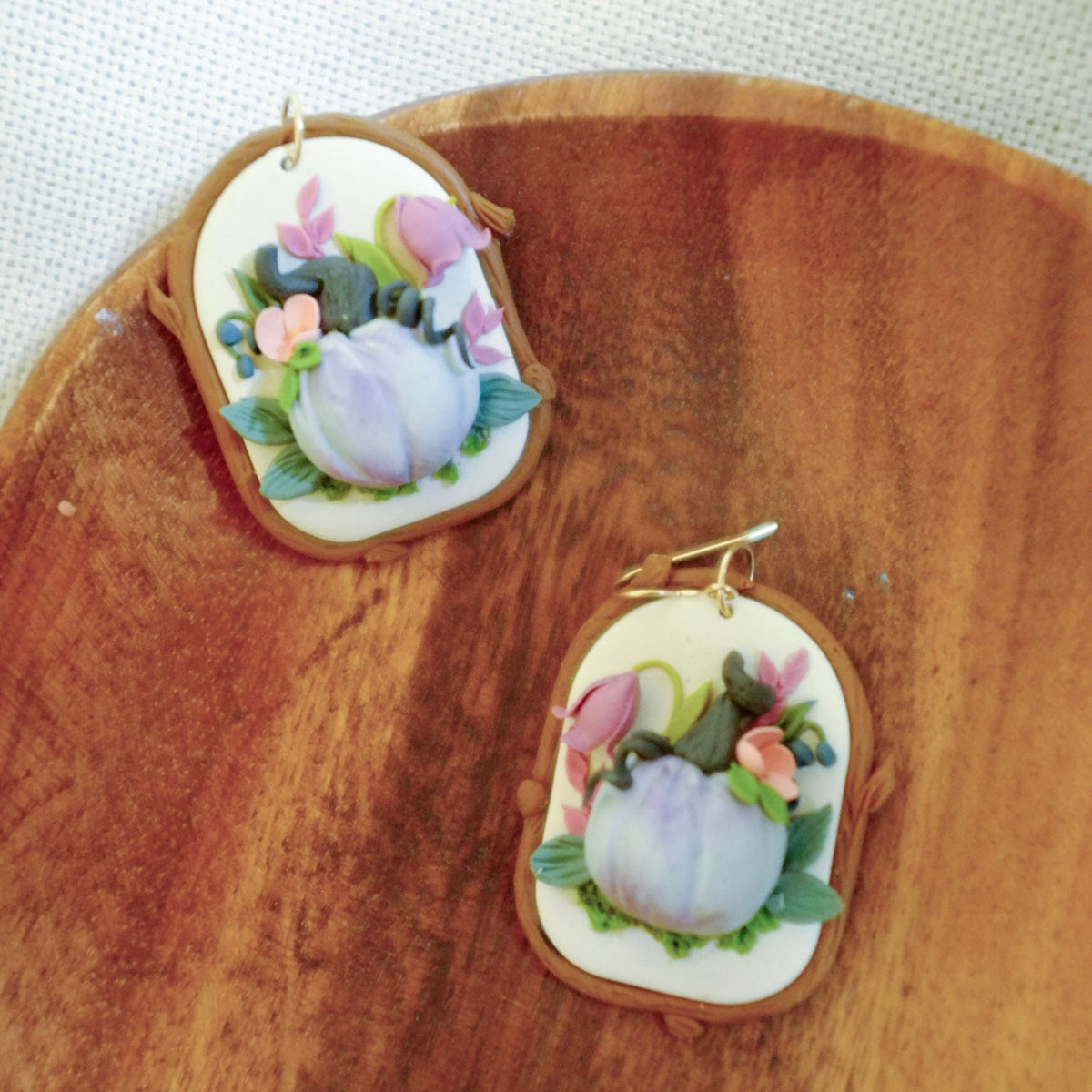 A pair of oval earrings with hand-painted floral designs featuring lavender, pink, and green on a cream background, set in a gold-tone frame and displayed on a wooden surface.