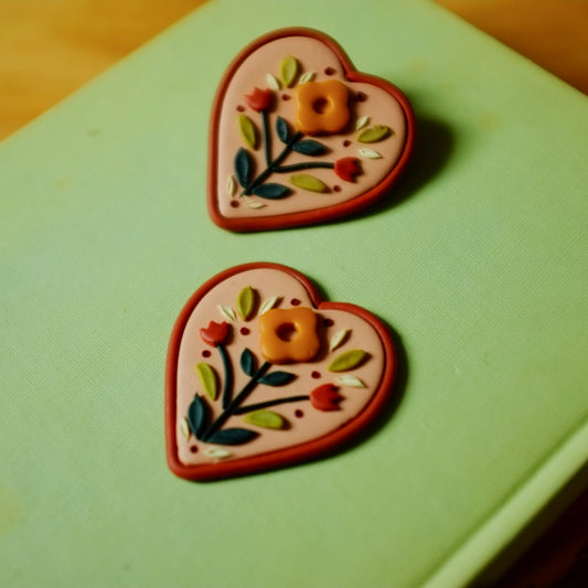 Folk style and heart shaped clay floral earrings with warm terra cotta colors, set on a light green backdrop