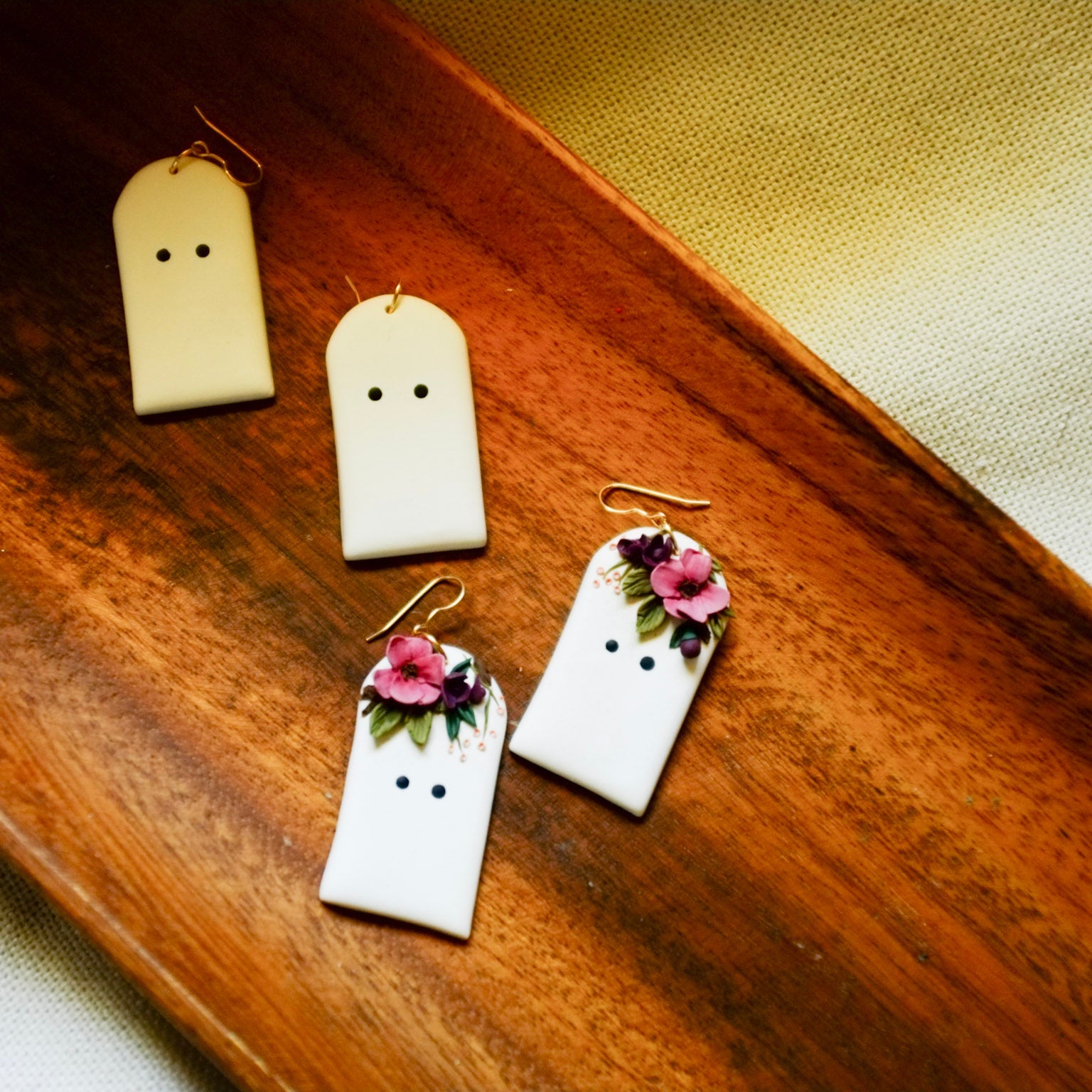Four ghost-shaped earrings displayed on a wooden tray, featuring two plain cream-colored earrings and two white earrings decorated with pink flowers and green leaves.