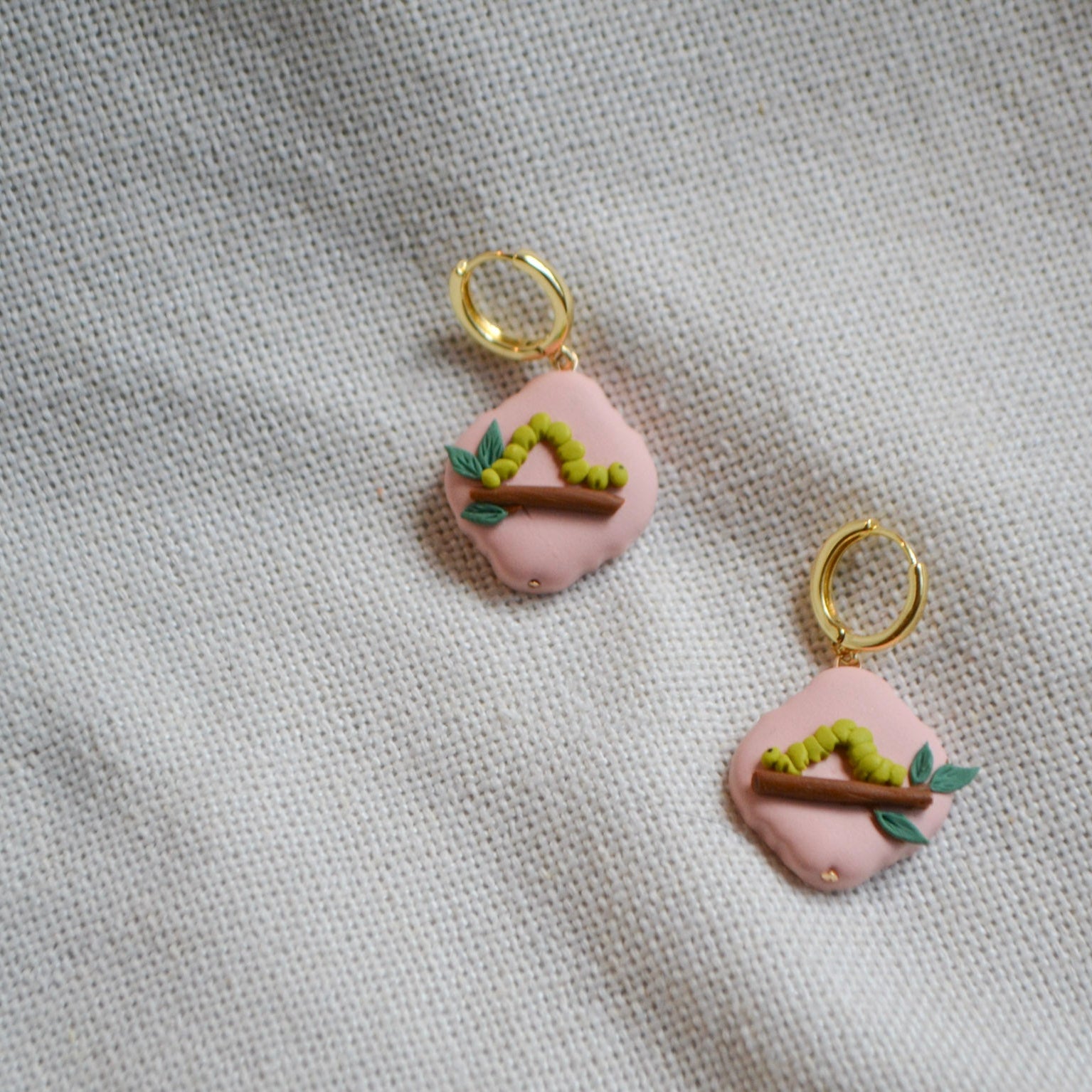 a pair of earrings with an inchworm on a leafy branch on a peach background with gold hoops