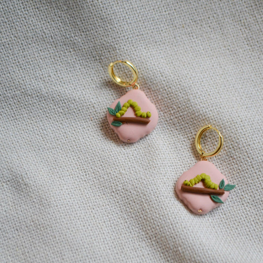 a pair of earrings with an inchworm on a leafy branch on a peach background with gold hoops