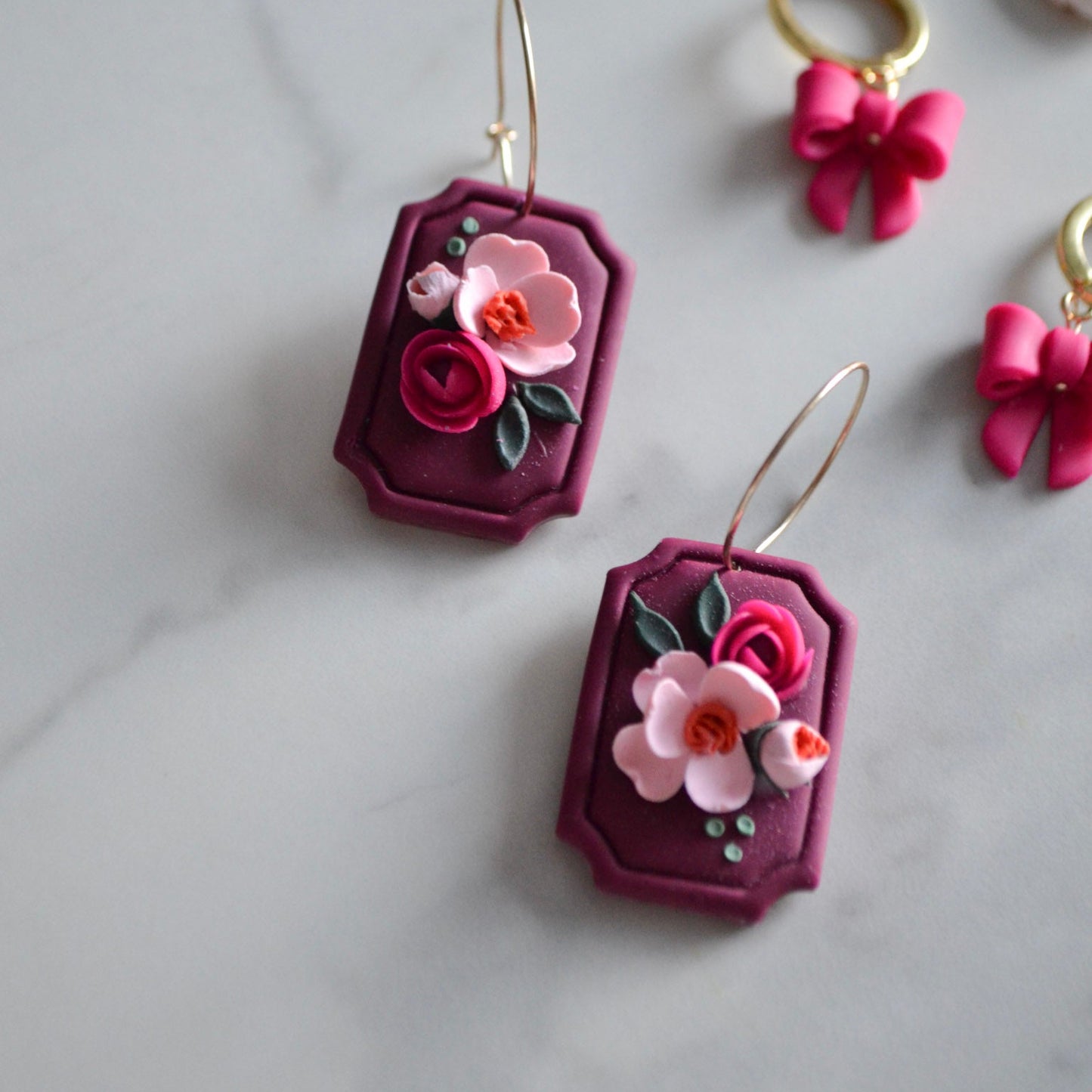 A pair of maroon earrings with gold-filled hoops, featuring pink floral designs and green accents, displayed on a surface with other related jewelry items.