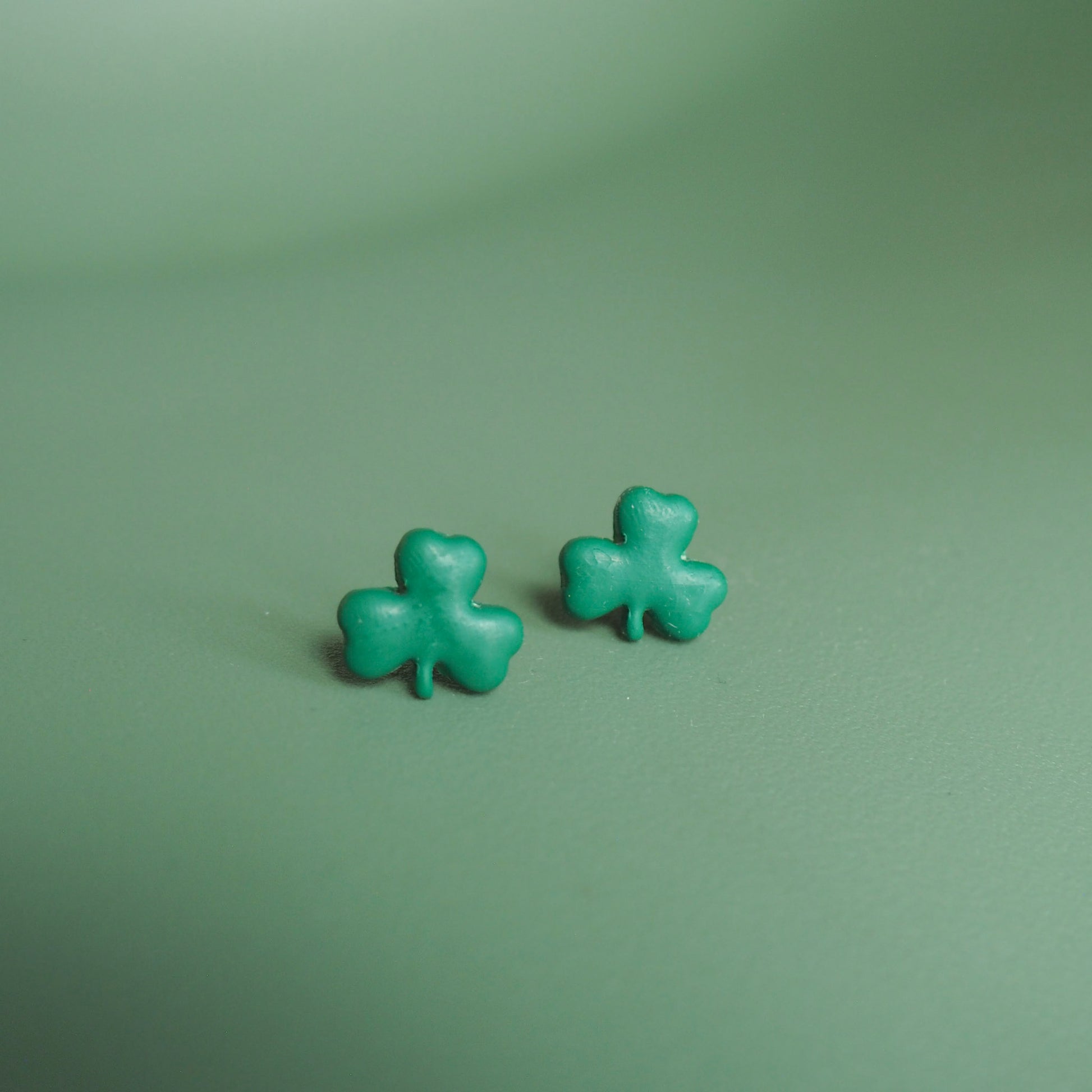 Small green shamrock polymer clay stud set on a lighter green background. 